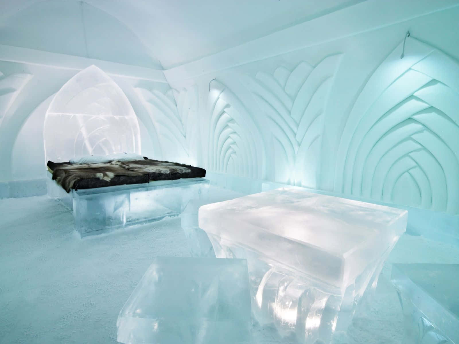 Ice Hotel 桌布 壁紙 — 寧靜的冰酒店房間，雕刻的拱形牆壁在柔和的藍光下閃耀，床上鋪著深色毛皮。