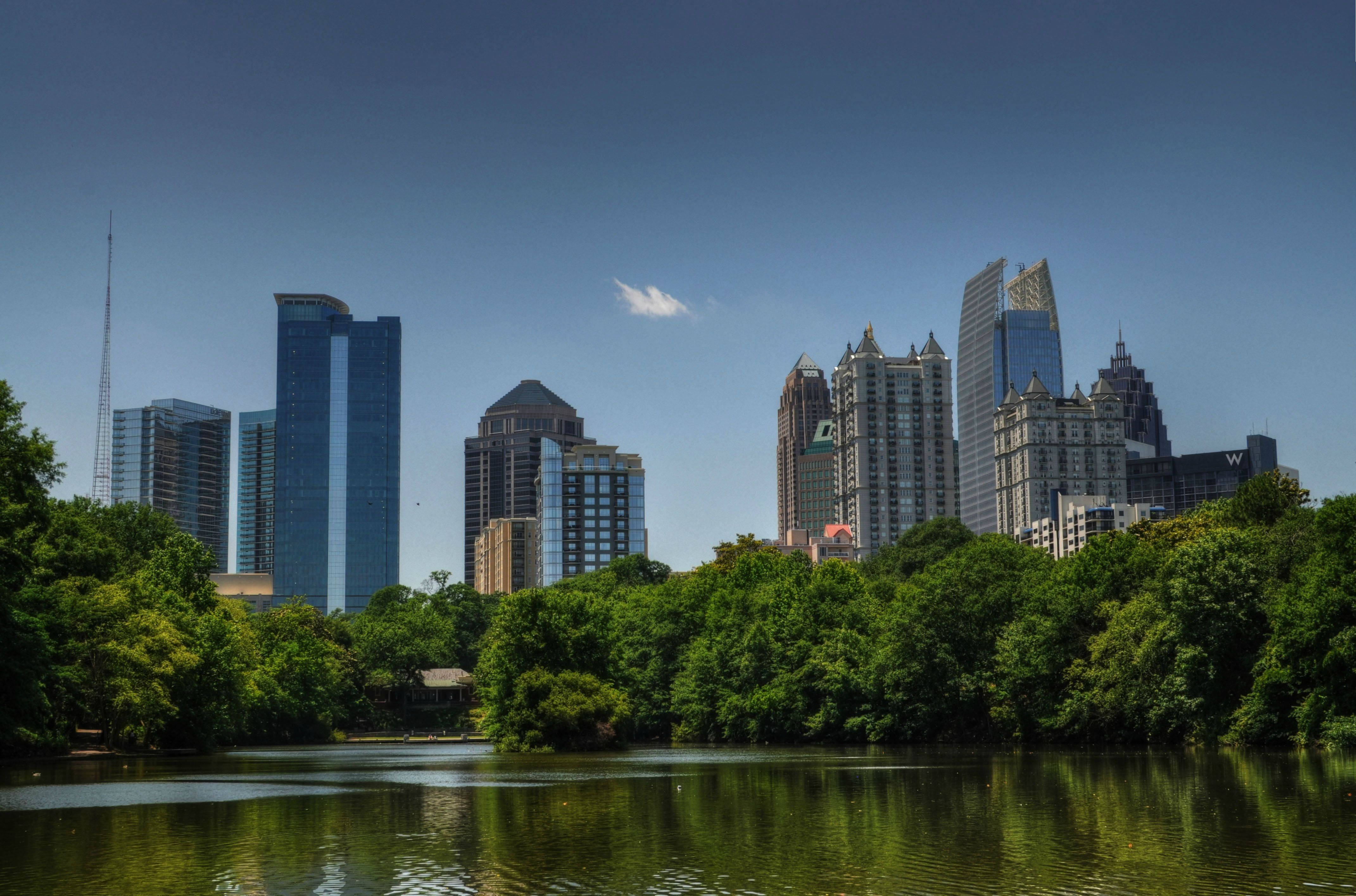 Atlanta 桌布 壁紙 — 寧靜的城市天際線，現代摩天大樓倒映在平靜的湖面上，周圍環繞著茂密的綠樹，天空清澈湛藍。