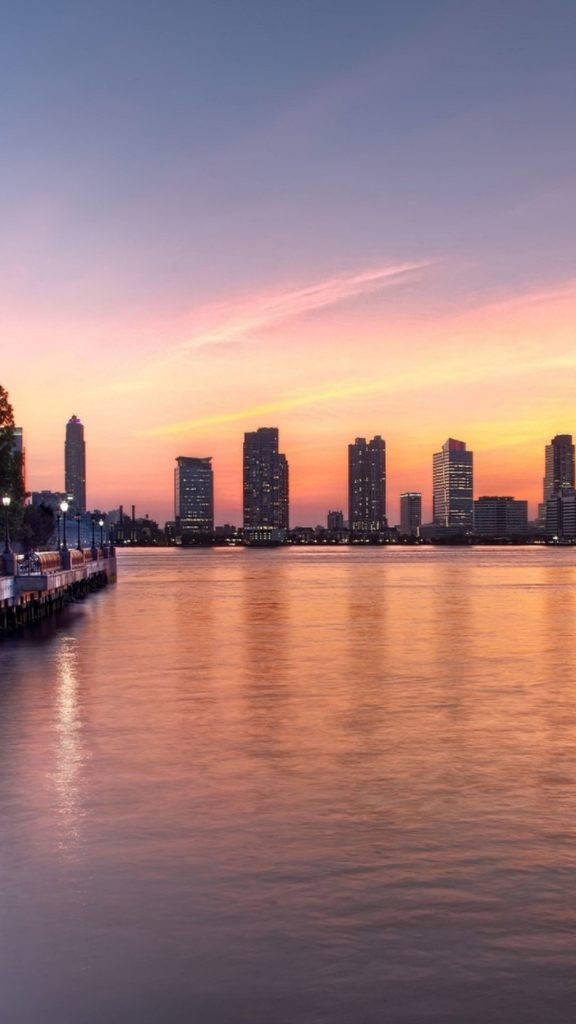 New 桌布 壁紙 — 寧靜的城市天際線在日落時分，高樓大廈在平靜的水面上反射出溫暖的橙色和粉紅色調。