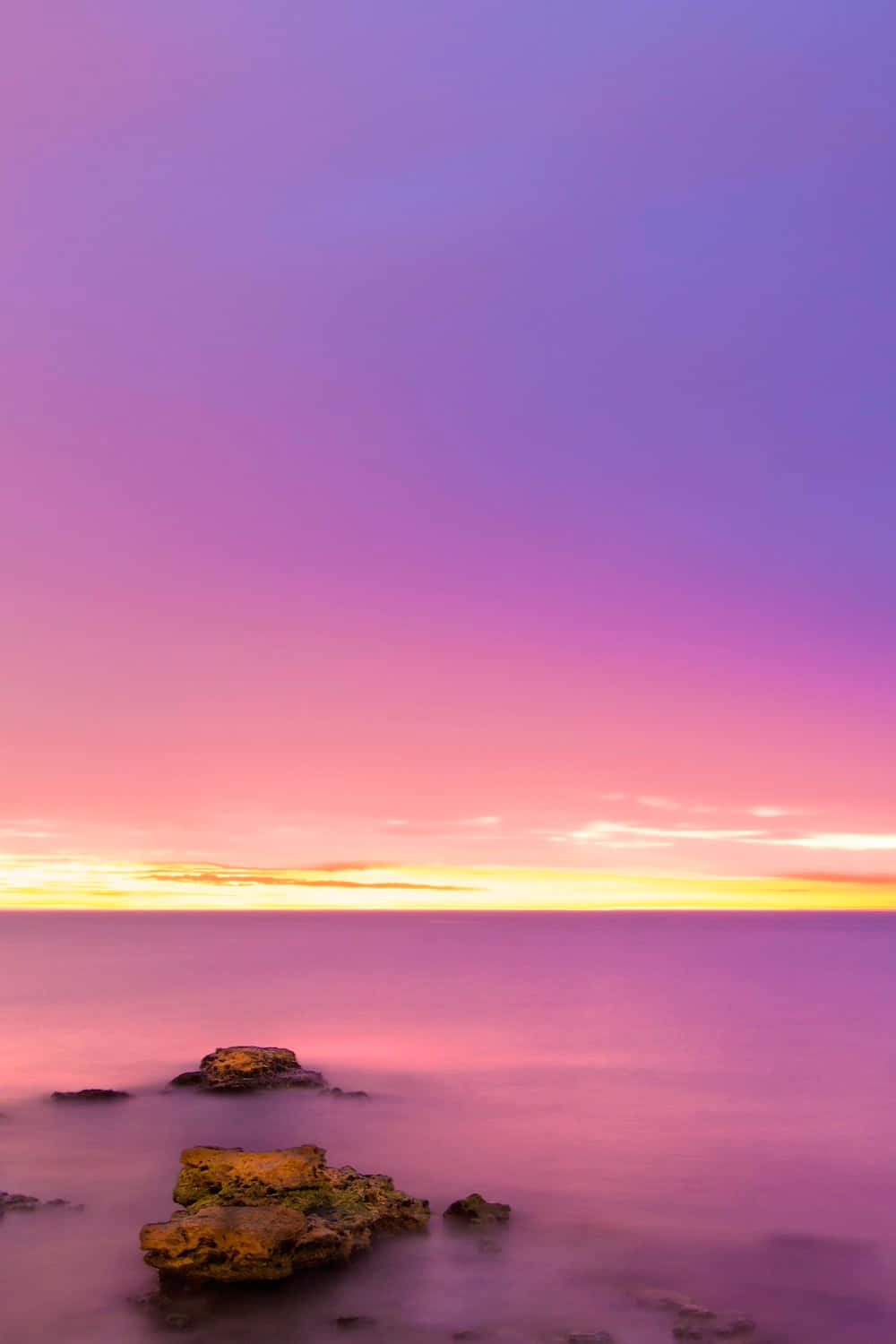 Pink And Yellow 桌布 壁紙 — 寧靜的夕陽在平靜的海面上描繪出充滿活力的紫色、粉紅色和金色的天空，前景有岩石的剪影。