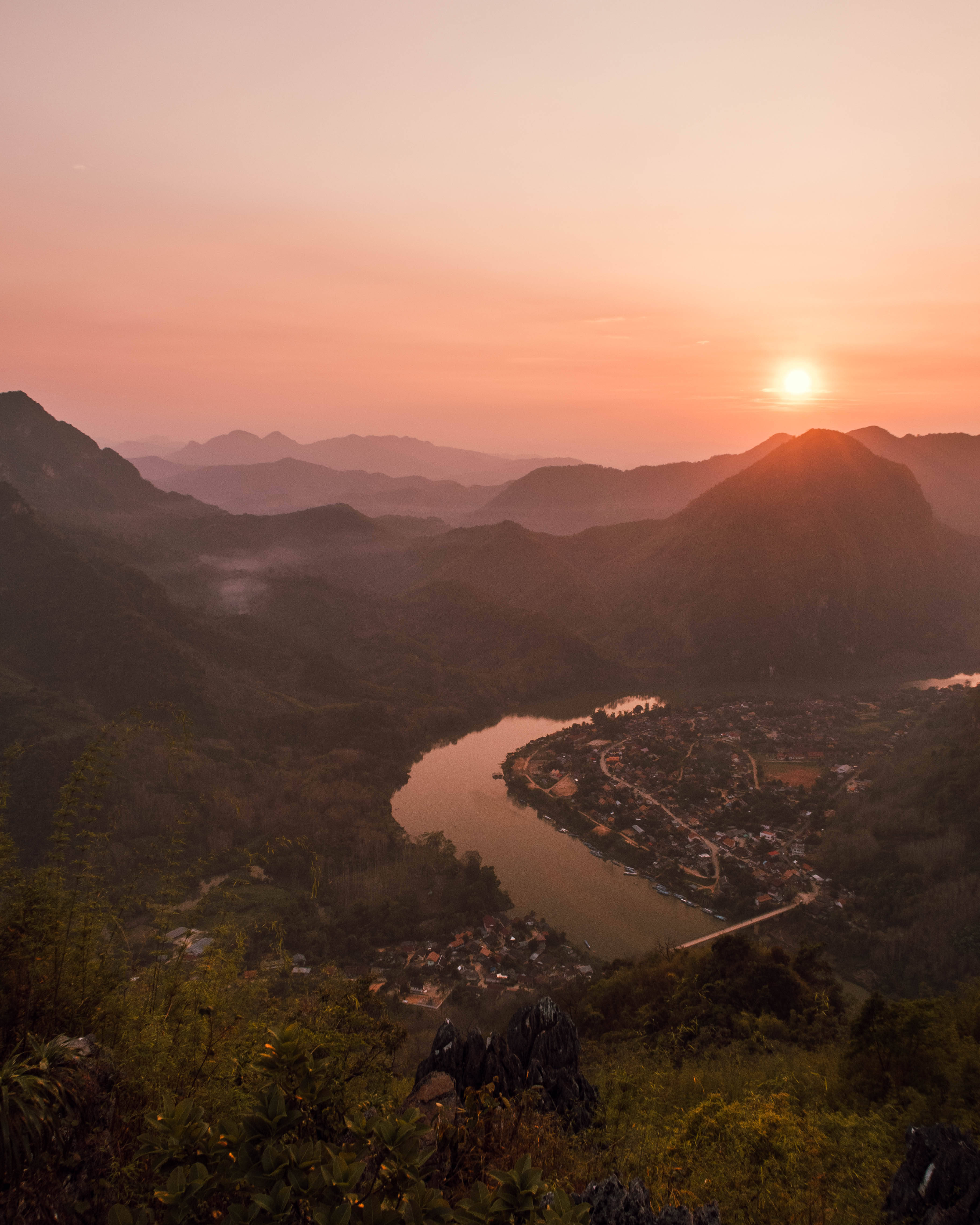 Laos 桌布 壁紙 — 寧靜的夕陽將蜿蜒的河流和依偎的村莊沐浴在溫暖的橙色和粉紅色調中，周圍是霧氣繚繞的山巒。