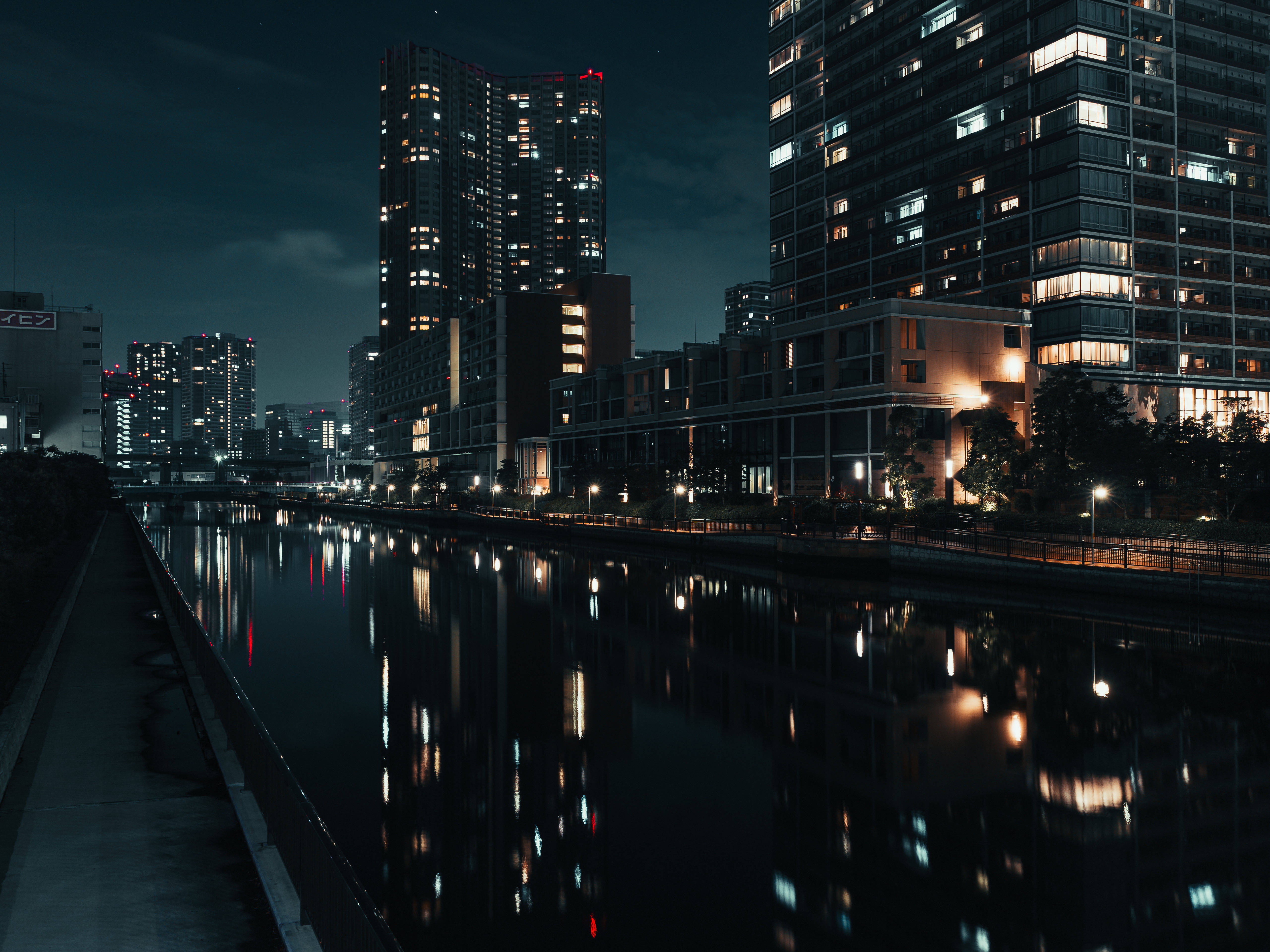東京 桌布 壁紙 — 寧靜的夜晚城市景觀，高樓大廈在黑暗的天空下將溫暖的燈光反射在平靜的河面上。