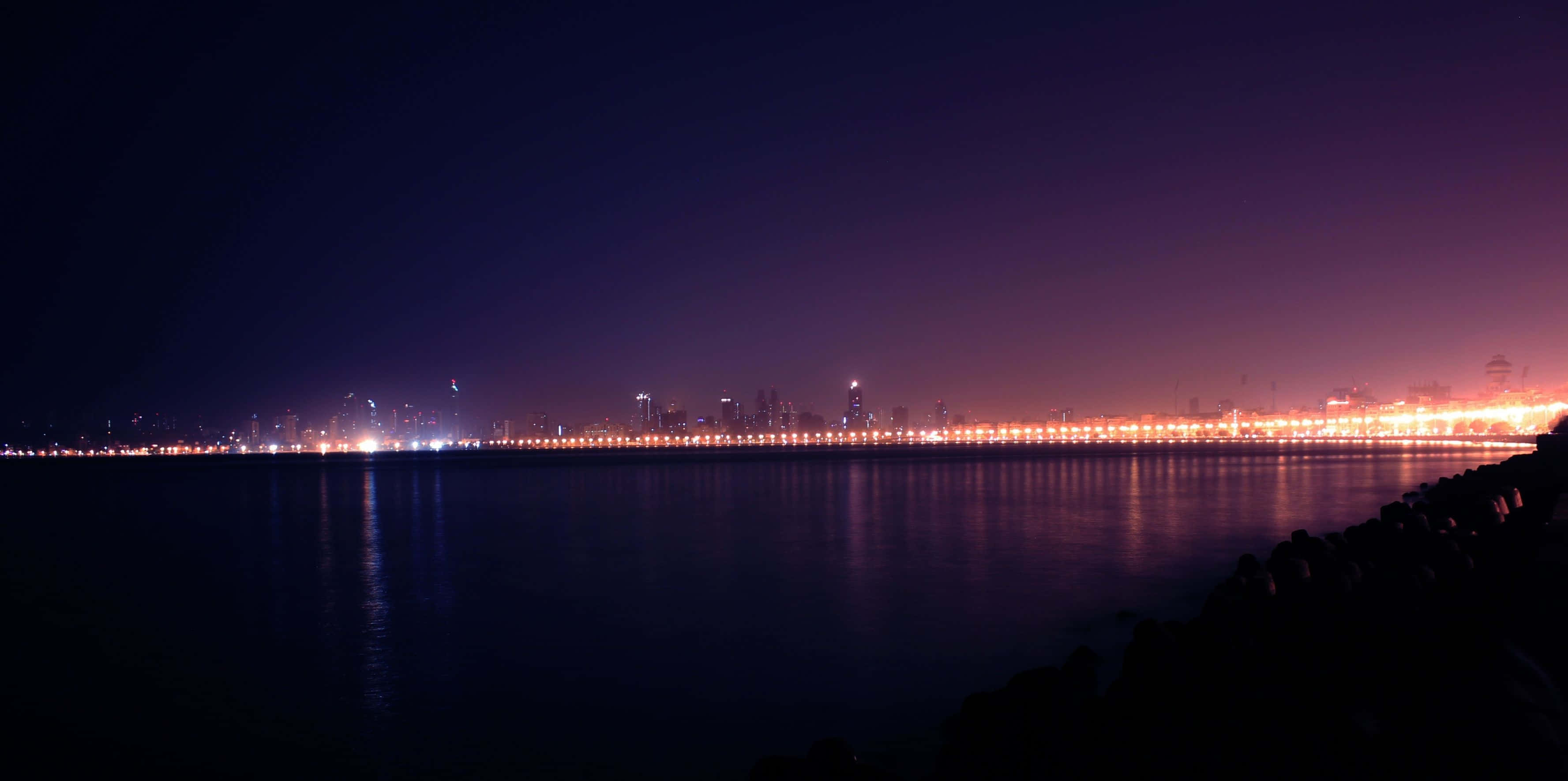 Mumbai 桌布 壁紙 — 寧靜的夜晚景色，城市天際線倒映在平靜的水面上，帶有紫色和橙色的色調。