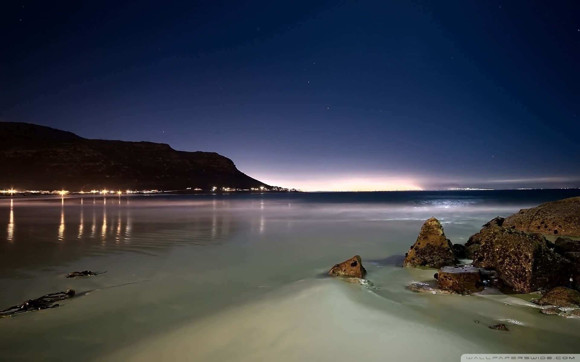 1920x1080 HD Beach 桌布 壁紙 — 寧靜的夜晚海灘場景，星空、遠處燈光映照在平靜的水面上，前景是崎嶇的岩石。