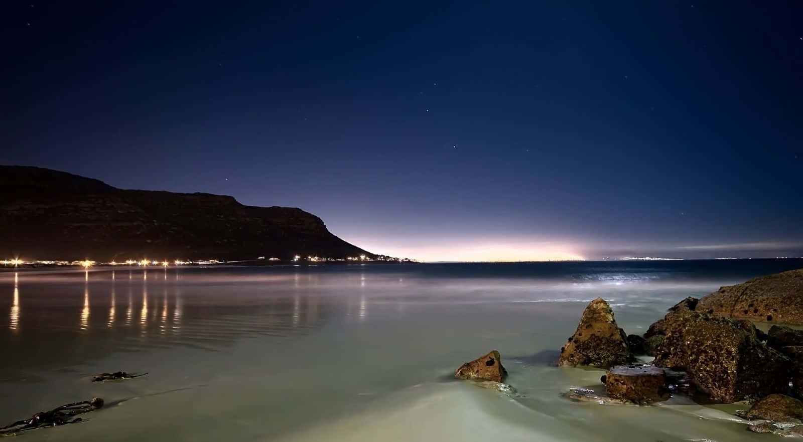 海灘桌面 桌布 壁紙 — 寧靜的夜晚海灘場景，有星空、發光的地平線和平靜的水面，反射遠處的城市燈光。