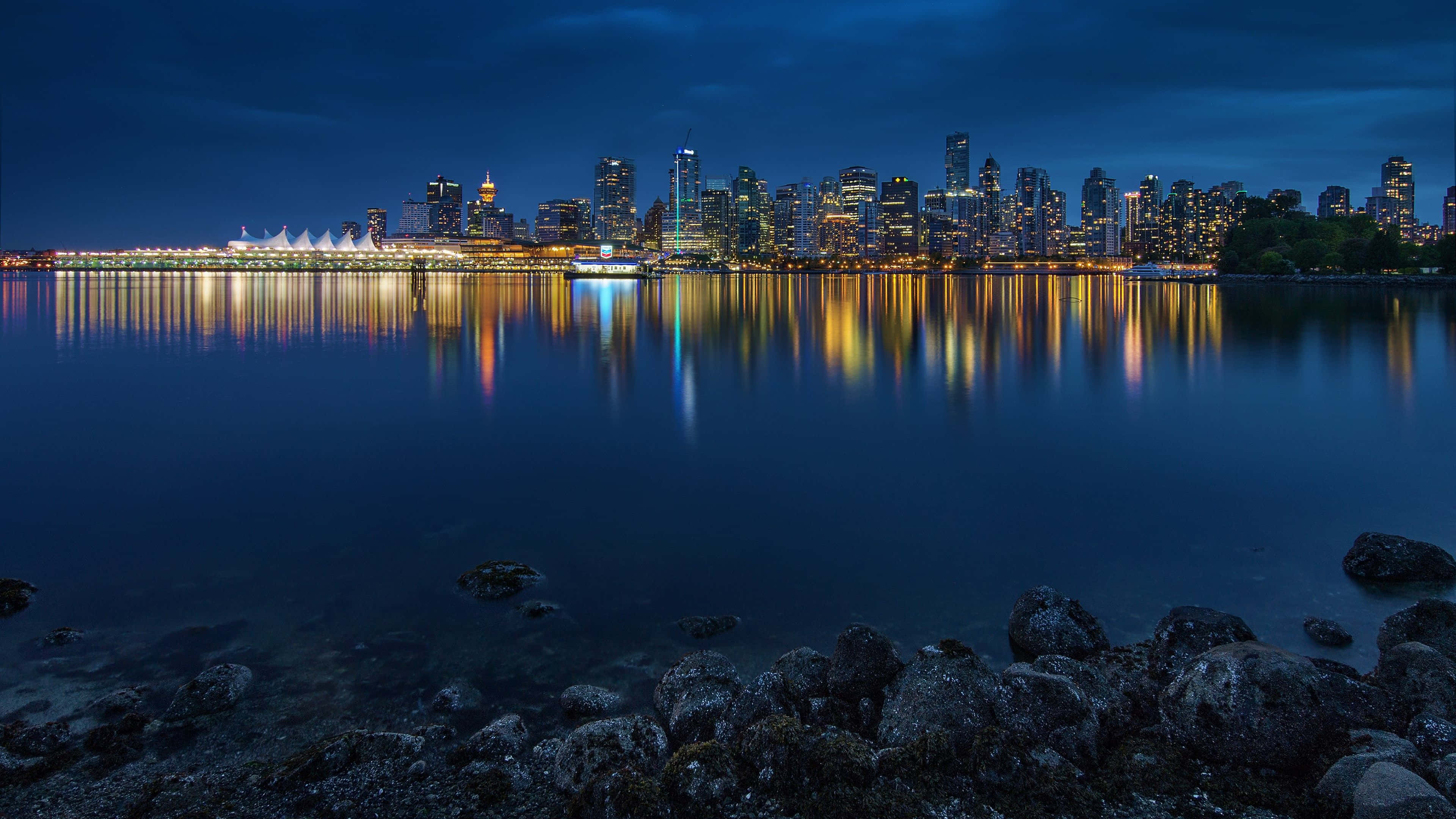 桌布 壁紙 — 寧靜的夜景，城市天際線倒映在平靜的水面上，前景岩石嶙峋。