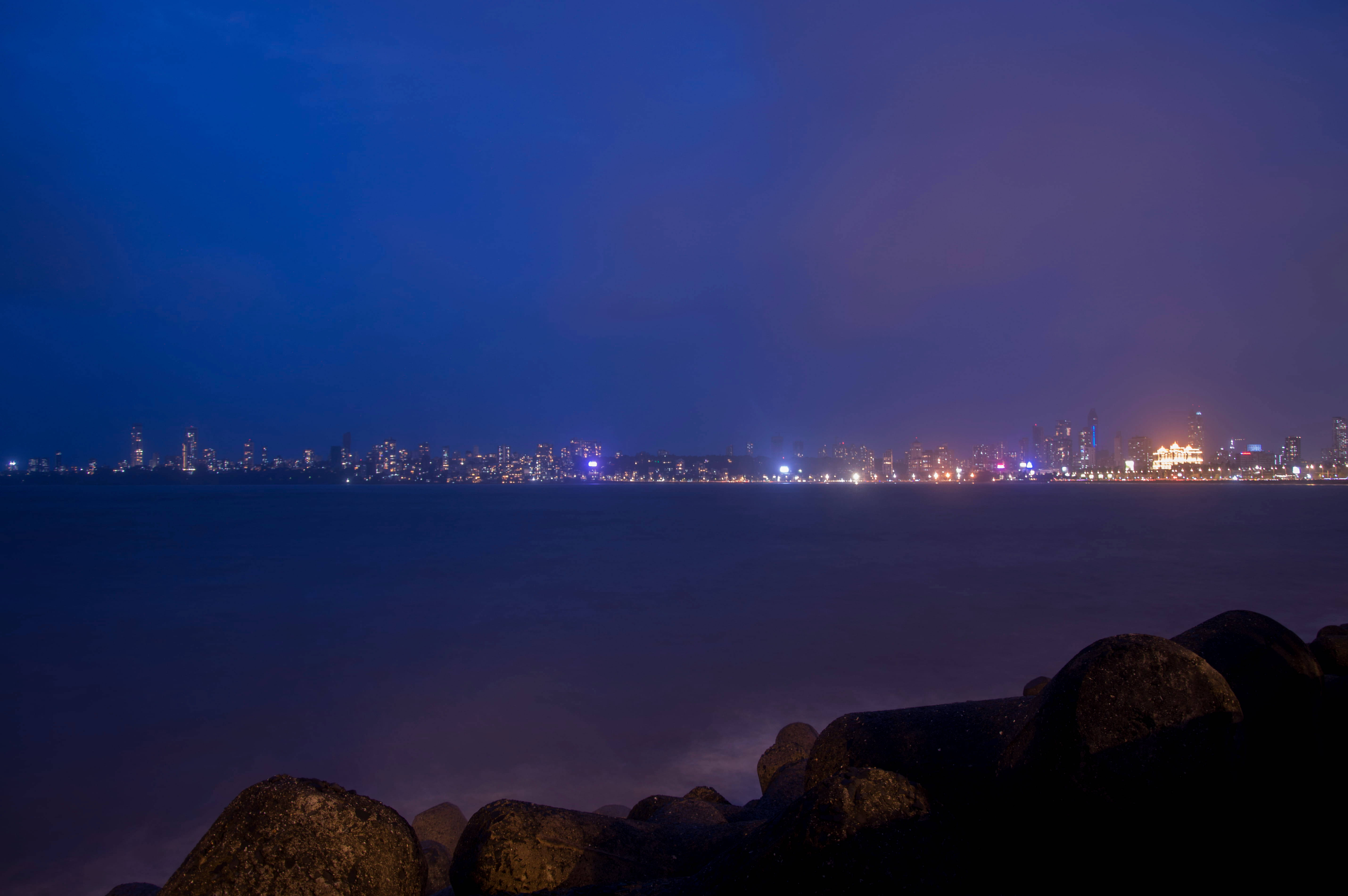Mumbai 桌布 壁紙 — 寧靜的夜景，城市天際線在平靜的海面上閃耀，前景有深色岩石。