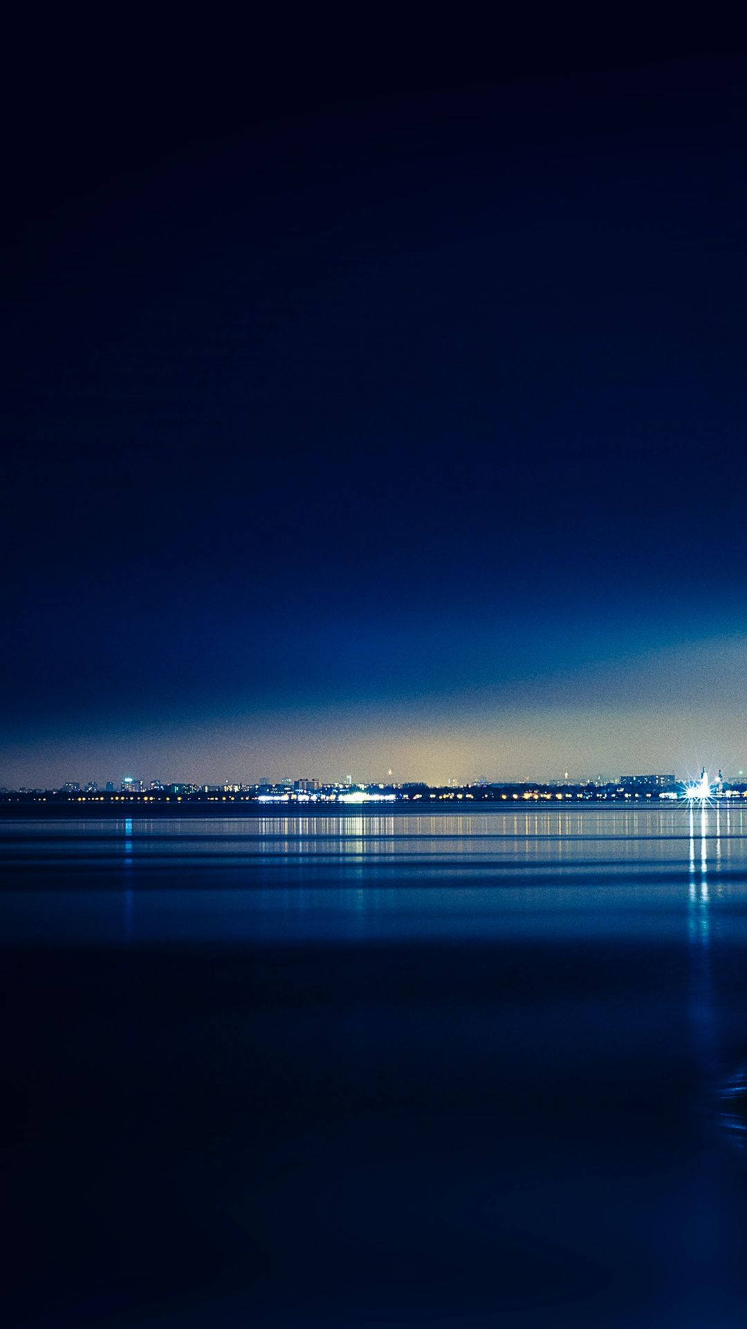 深藍色美學動圖 桌布 壁紙 — 寧靜的夜景，平靜的水面在深藍色天空下倒映著遠處的城市燈光。