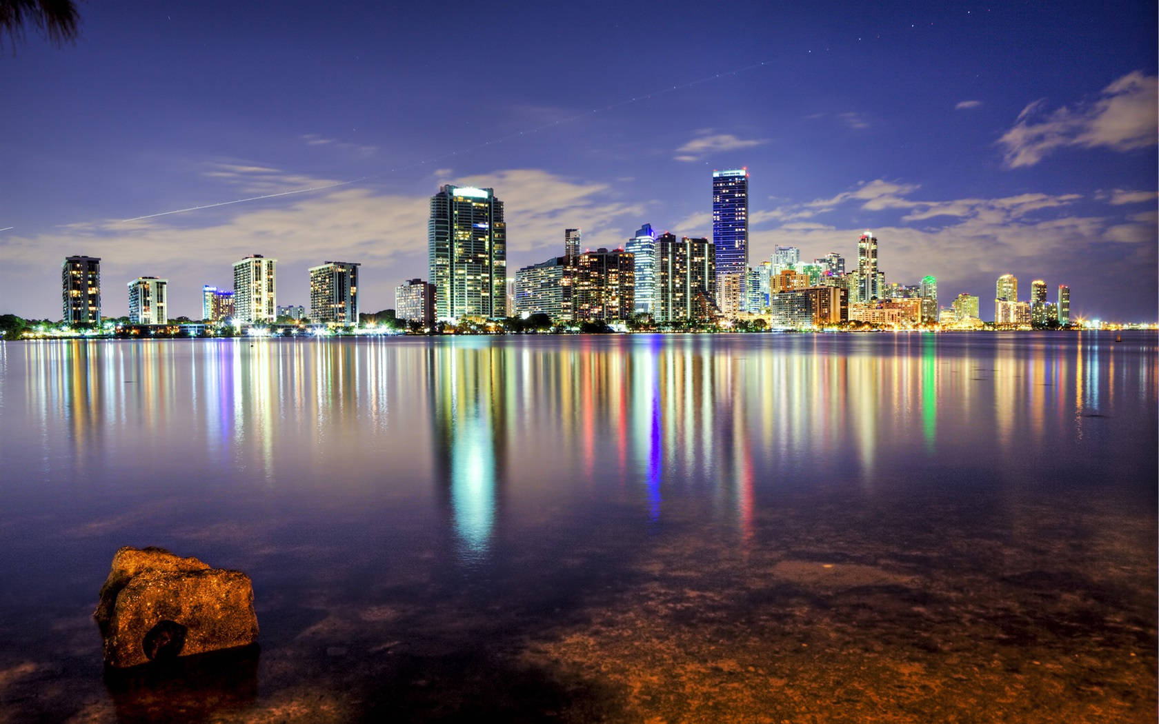 Orange City 桌布 壁紙 — 寧靜的夜景，摩天大樓在深藍色天空下閃耀，燈光完美倒映在平靜的水面上，前景有一塊孤獨的岩石。