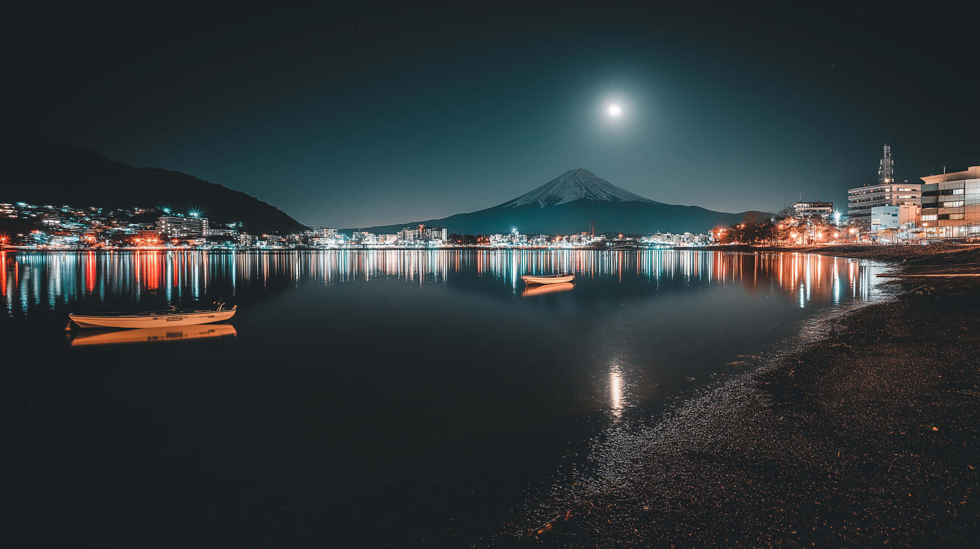 Mount Fuji Reflected in Lake with City Lights at Night 壁紙 — 寧靜的夜景，滿月照亮富士山及其在平靜水面上的倒影，旁邊有一艘小船和城市燈光。