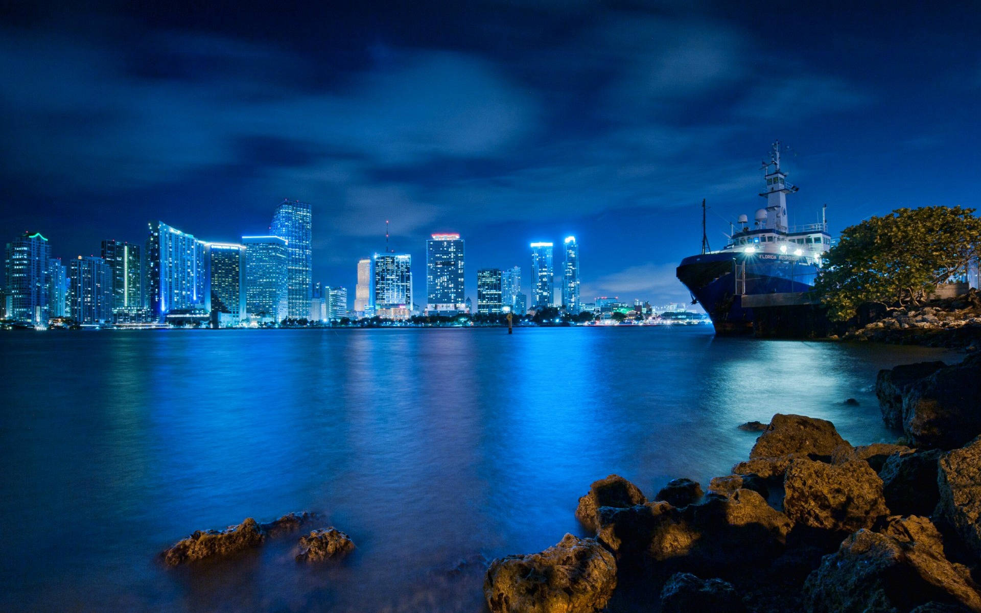 淺藍色 桌布 壁紙 — 寧靜的夜景，發光的城市天際線倒映在平靜的藍色海水中，旁邊是停泊的船隻和岩石海岸。