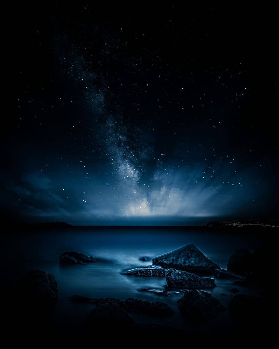 iPhone 桌布 壁紙 — 寧靜的夜間海景，發光的藍色海水映照著銀河和散落的星星，上方是黑暗的岩石。