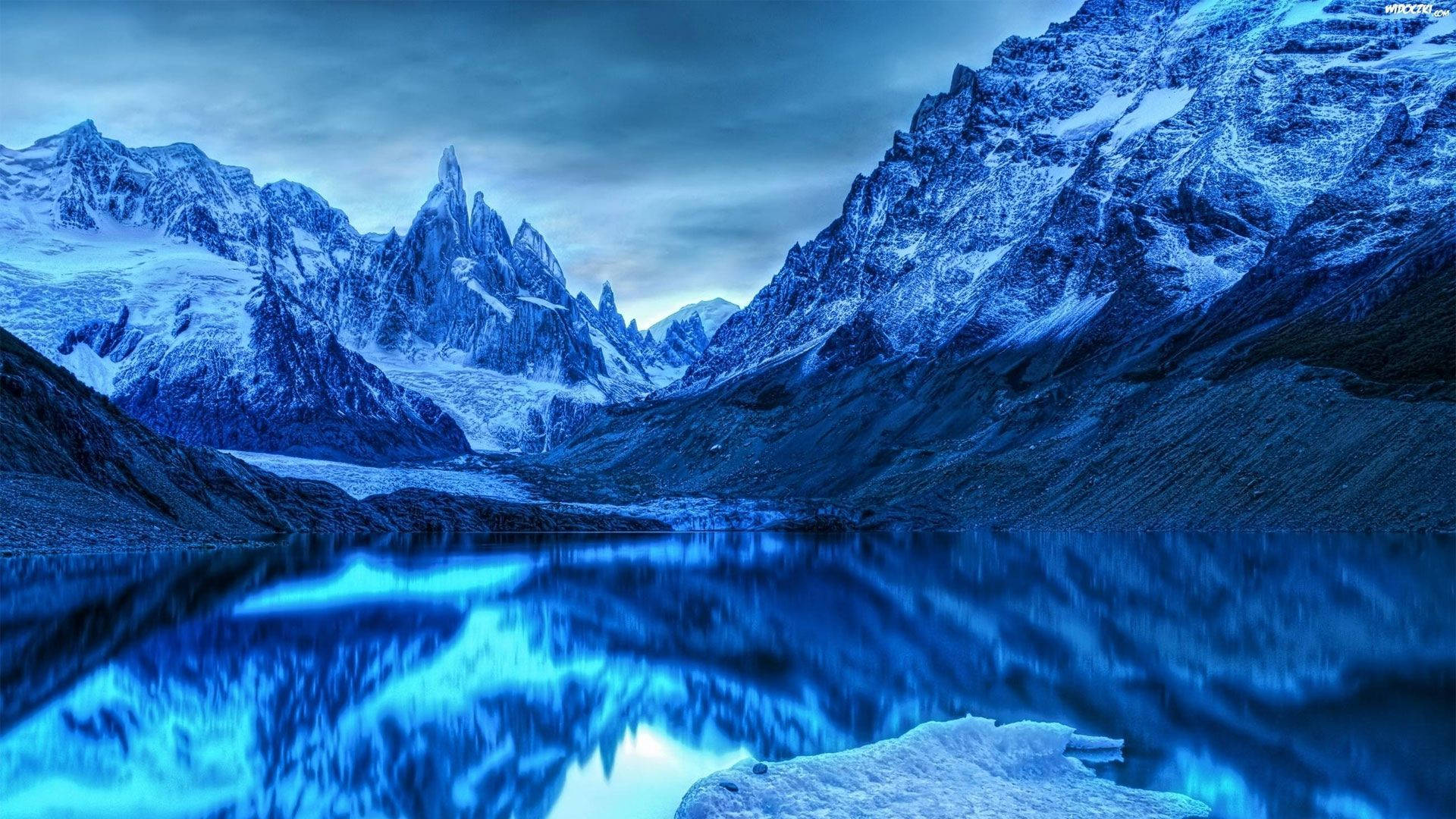 Patagonia 桌布 壁紙 — 寧靜的山湖在憂鬱的暮色天空下，以鮮豔的藍色調映照著積雪的山峰。