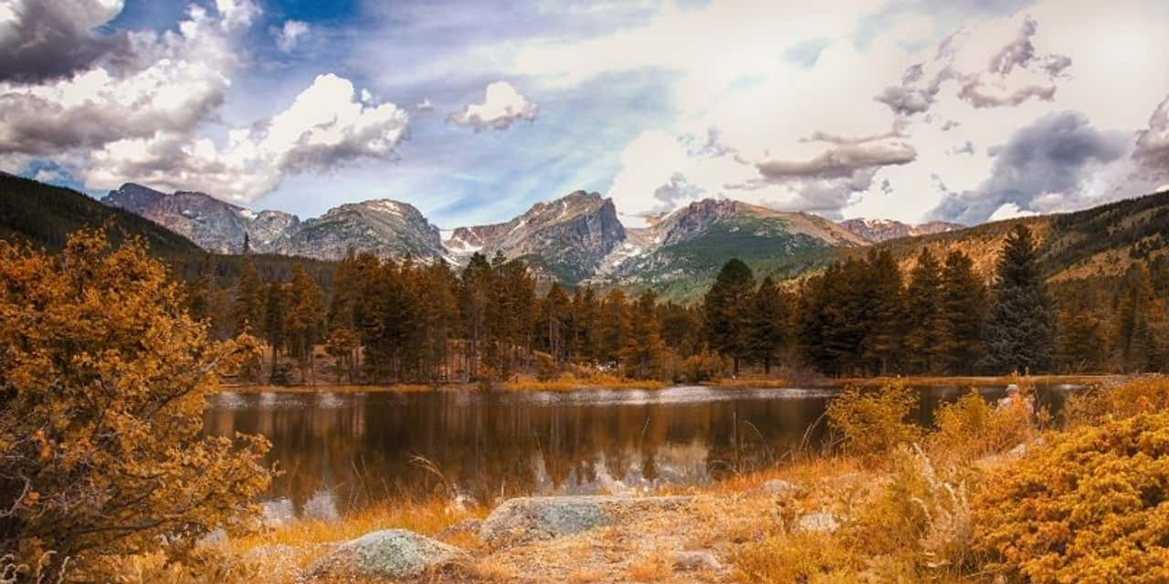 Yellowstone 桌布 壁紙 — 寧靜的山湖在戲劇性的多雲天空下倒映著積雪的山峰，前景點綴著秋葉。