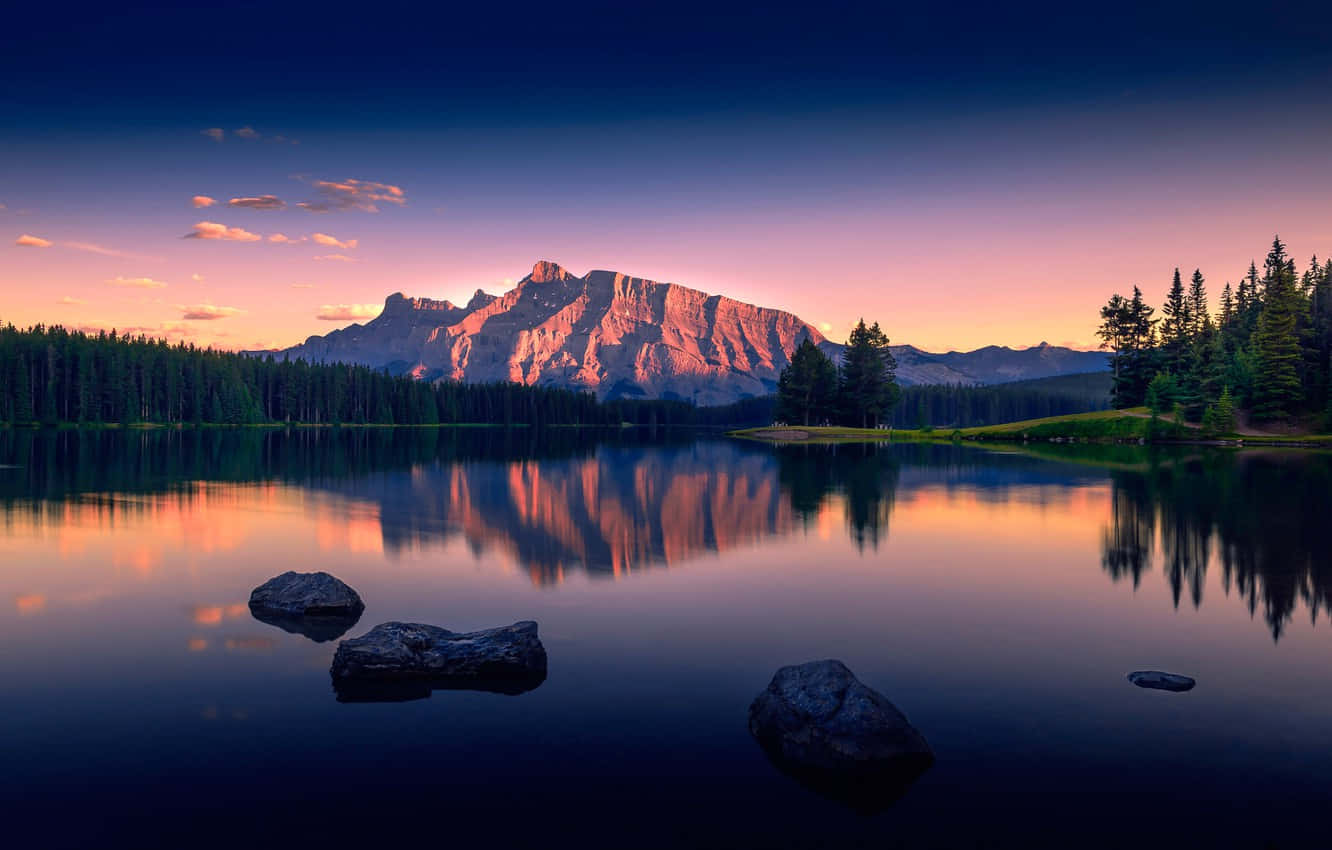 Serenity 桌布 壁紙 — 寧靜的山湖映照出夕陽西下時分，天空呈現粉紅與紫色調，映襯著崎嶇的山峰與常綠樹木。