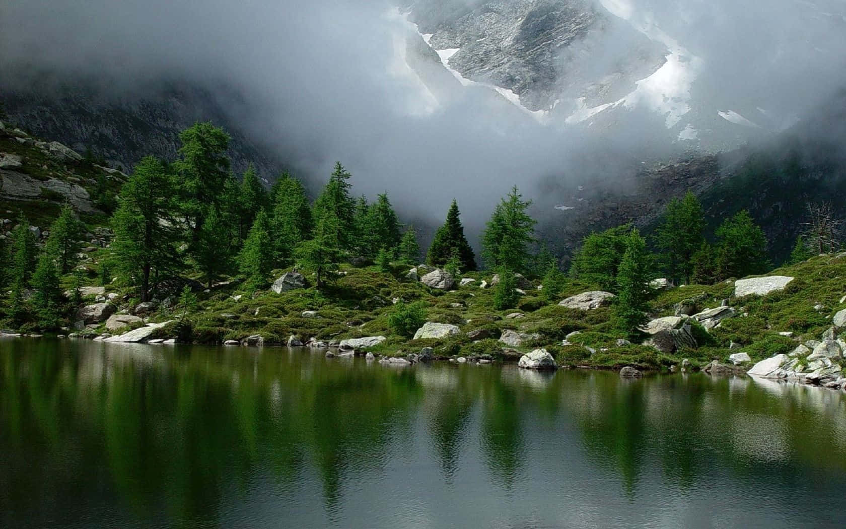 National Geographic Desktop 桌布 壁紙 — 寧靜的山湖映照著常綠樹和霧氣繚繞的山峰，上方聳立著積雪的山脊。