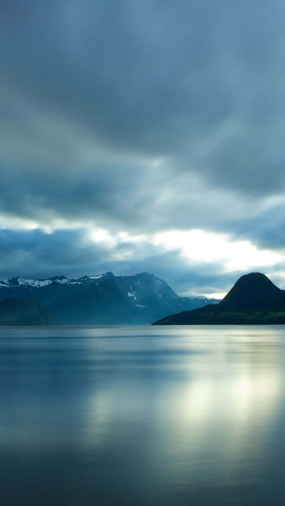Calming iPhone 桌布 壁紙 — 寧靜的山湖映照著遠方白雪覆蓋的山峰和戲劇性的多雲天空。