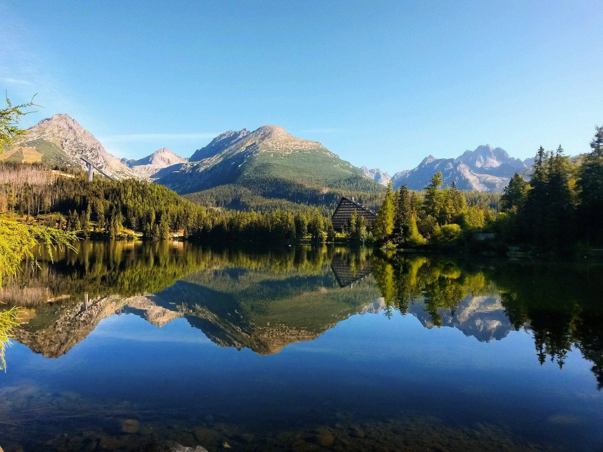 Slovakia 桌布 壁紙 — 寧靜的山湖映照著鋸齒狀的山峰和一座木屋，天空清澈湛藍。