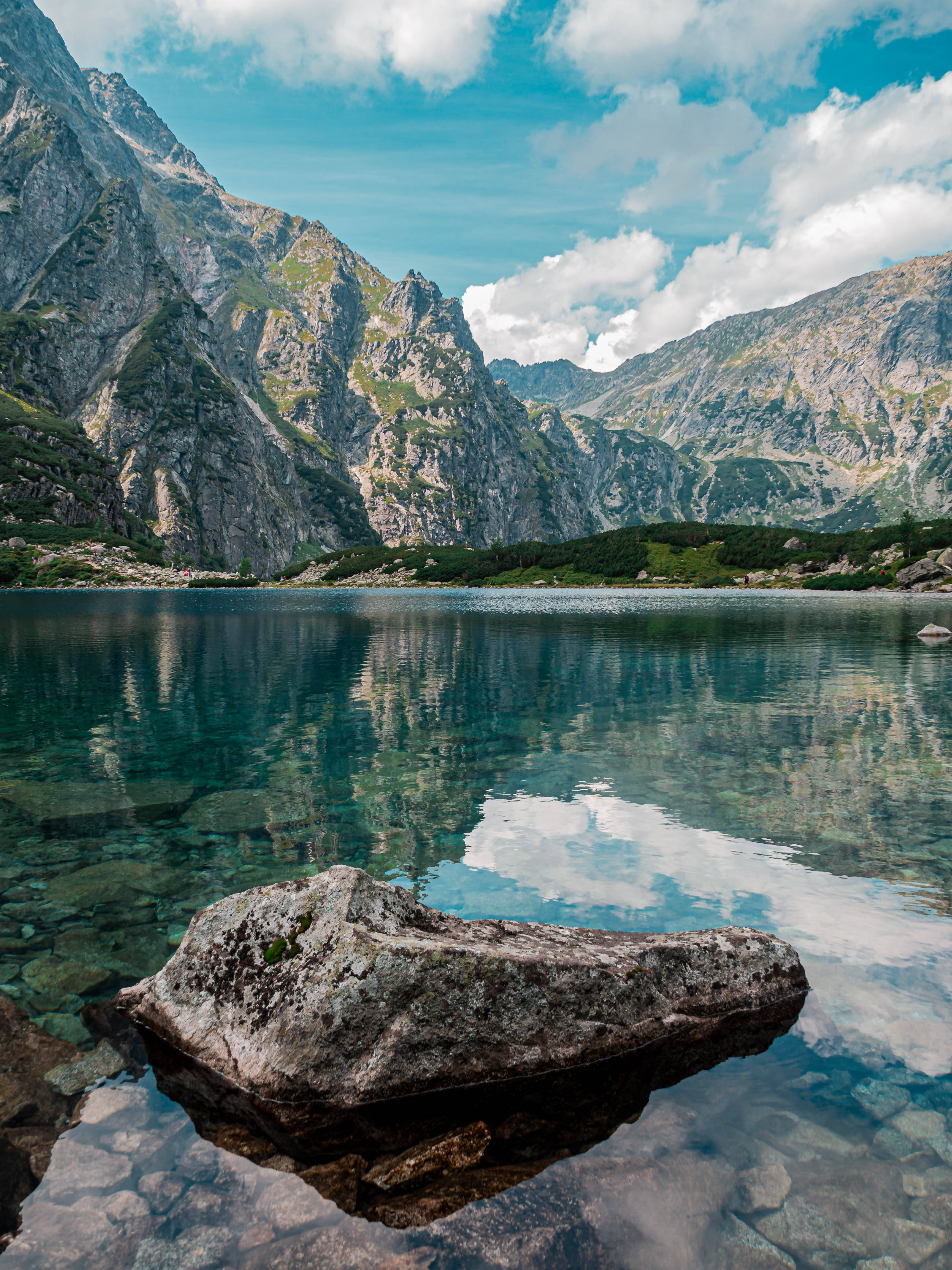 Lake 桌布 壁紙 — 寧靜的山湖映照著鋸齒狀的山峰和蓬鬆的雲朵，在明亮的藍天下，前景有一塊大岩石。
