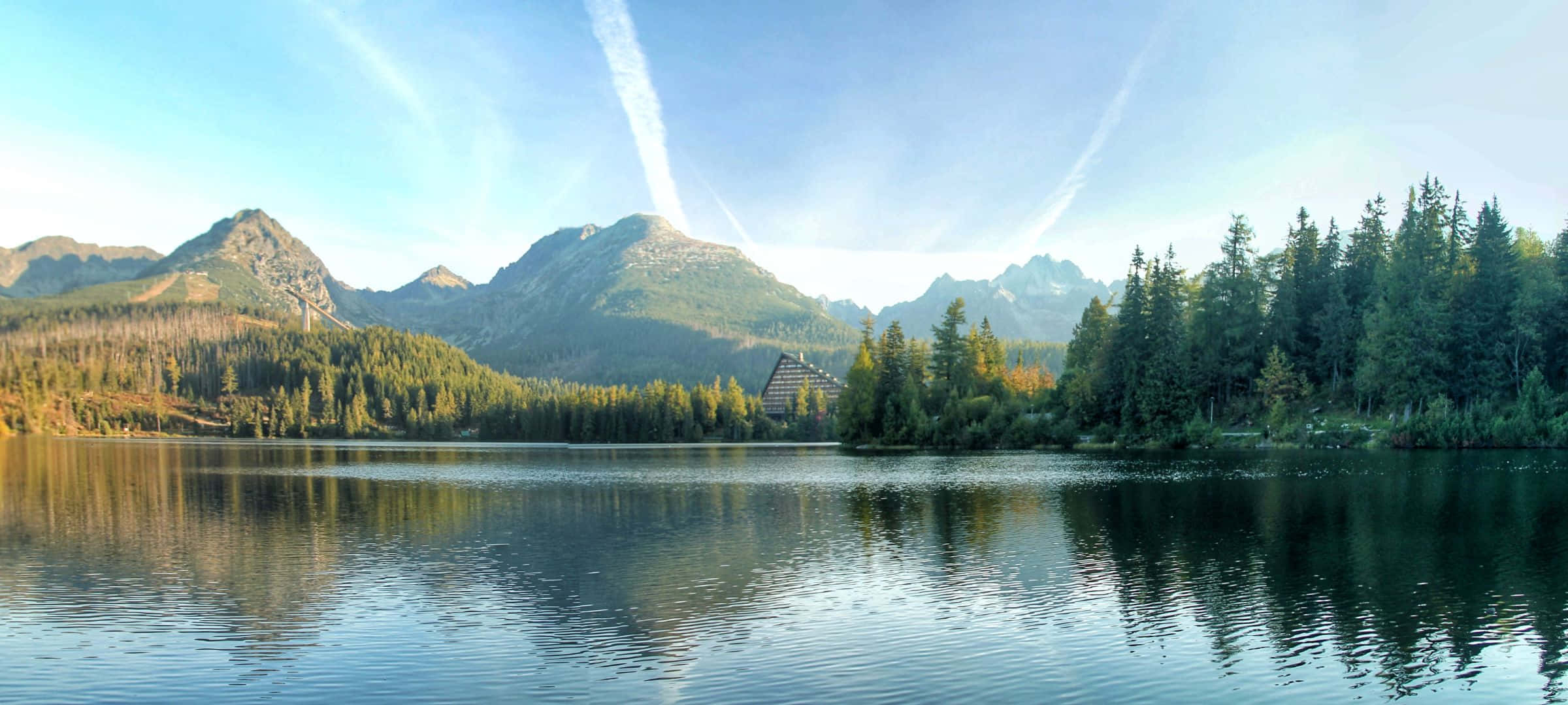 2400x1080 桌布 壁紙 — 寧靜的山間湖泊倒映著高聳的山峰和常綠森林，天空湛藍，雲朵輕柔。