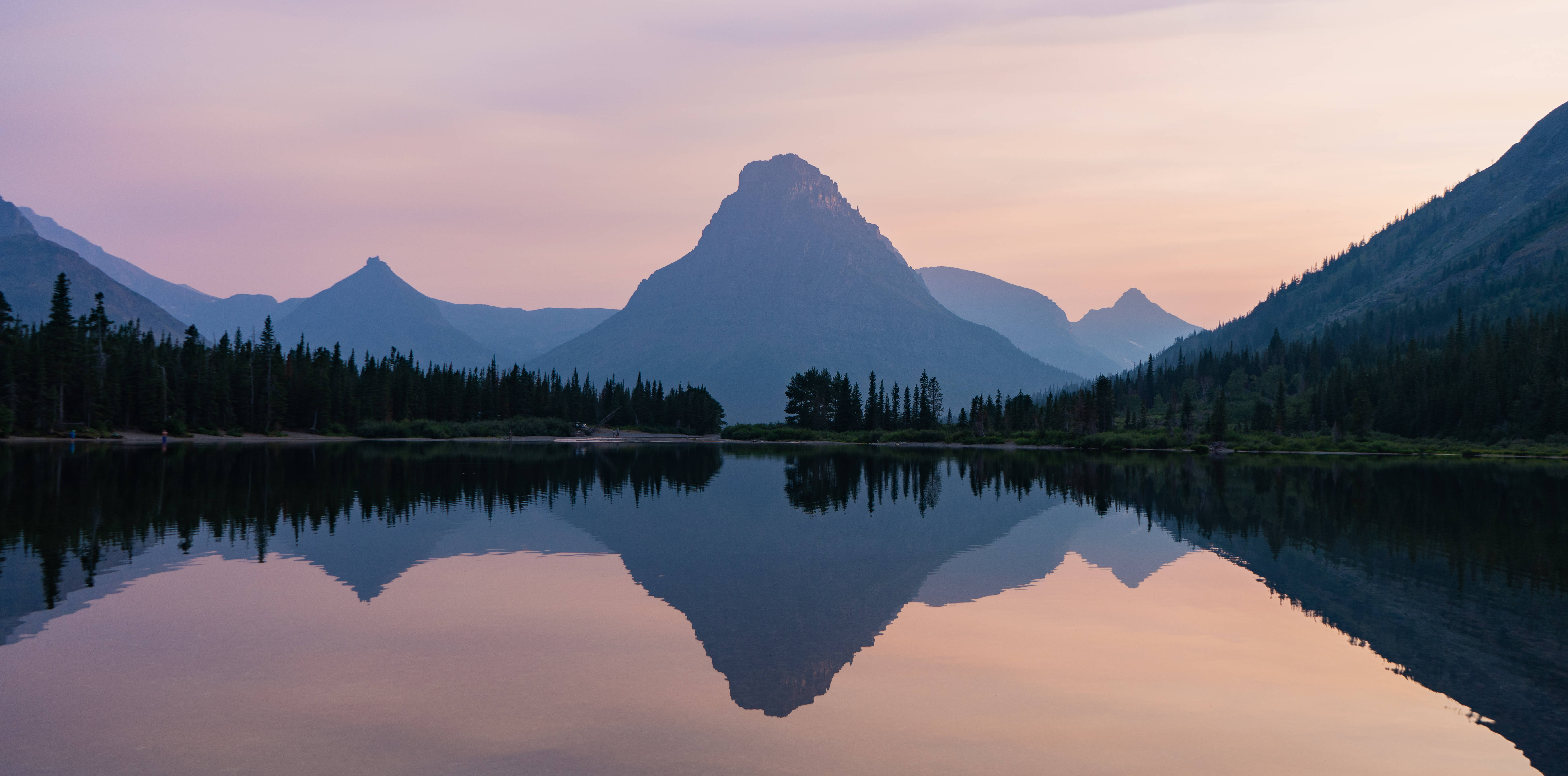 Montana 桌布 壁紙 — 寧靜的山間湖泊映照著粉紅與紫色的日落天空，襯以尖銳的山峰和常綠樹木。