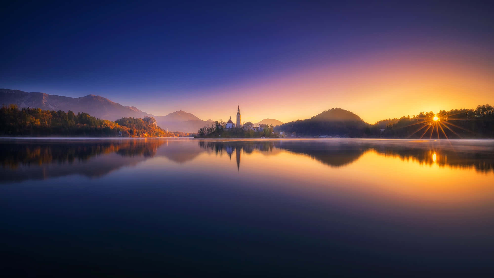 Lake Bled 桌布 壁紙 — 寧靜的日出，霧氣籠罩的湖面上，島上的教堂完美倒映在平靜的水面，四周環繞著秋日森林和遠山。
