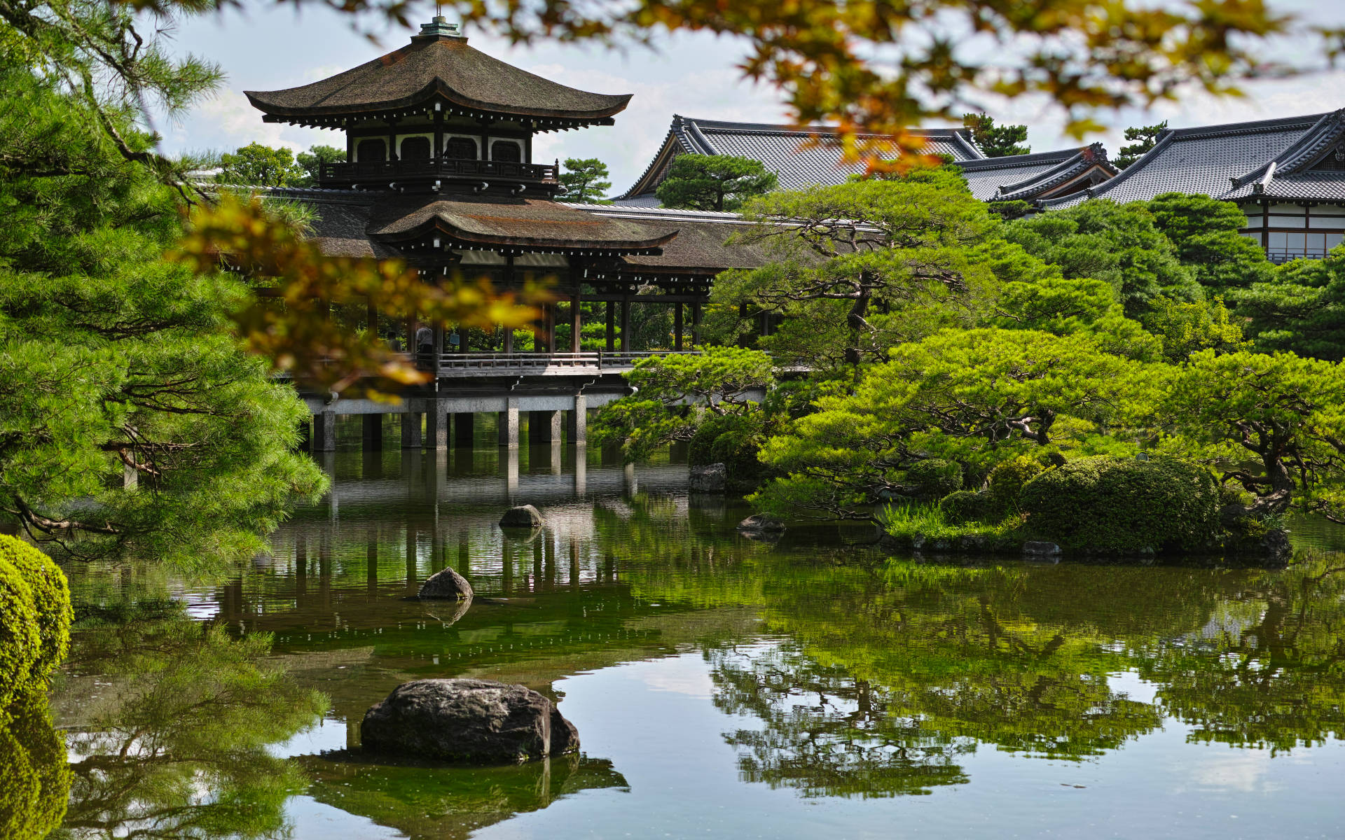 京都 桌布 壁紙 — 寧靜的日本庭園，傳統木造亭閣倒映在平靜的水面上，四周環繞著茂密的綠植和秋葉。