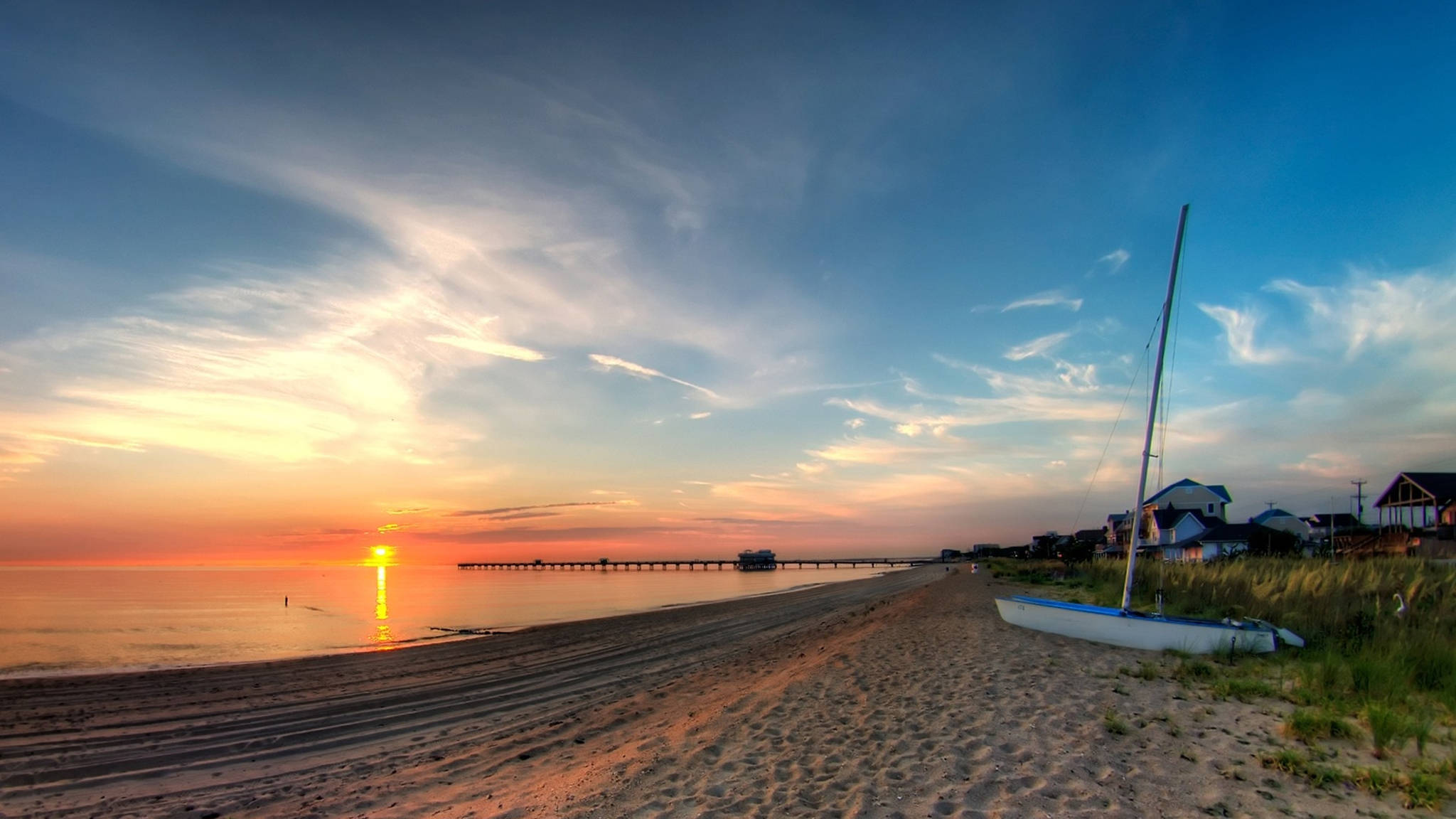 View 桌布 壁紙 — 寧靜的日落，平靜的海灘上有一艘帆船停泊在沙灘上，碼頭延伸至發光的水中。