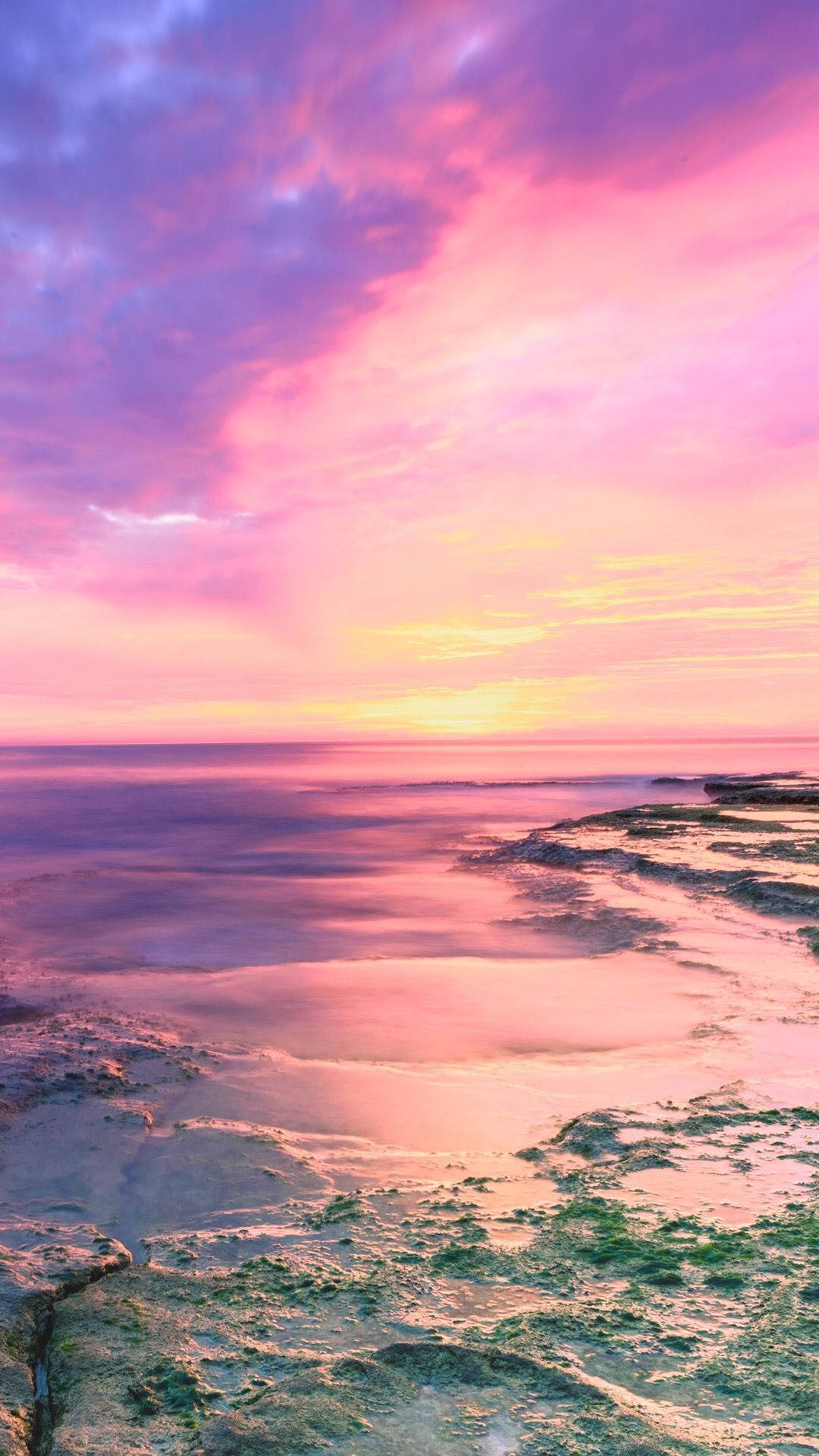Sunset Aesthetic 桌布 壁紙 — 寧靜的日落以鮮豔的粉紅色和紫色描繪天空，映照在平靜的海洋與岩石海岸交匯之處。