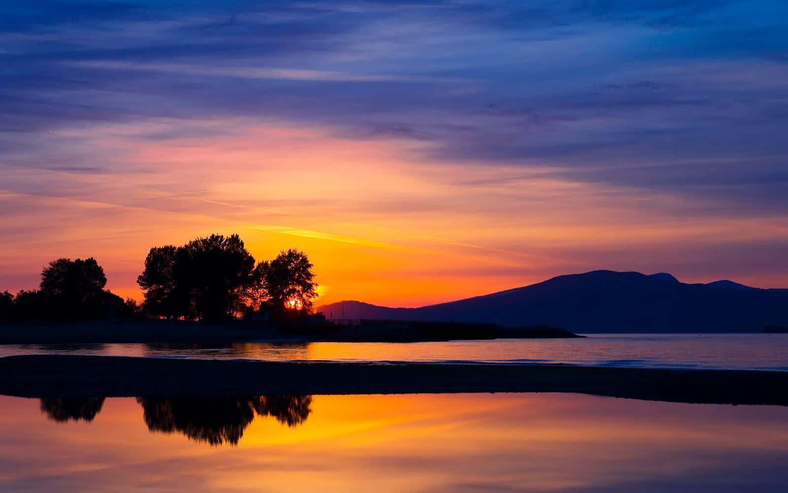Sunset Landscape 桌布 壁紙 — 寧靜的日落將天空染成火橙色和深紫色，樹木和山脈的剪影完美地倒映在平靜的水面上。