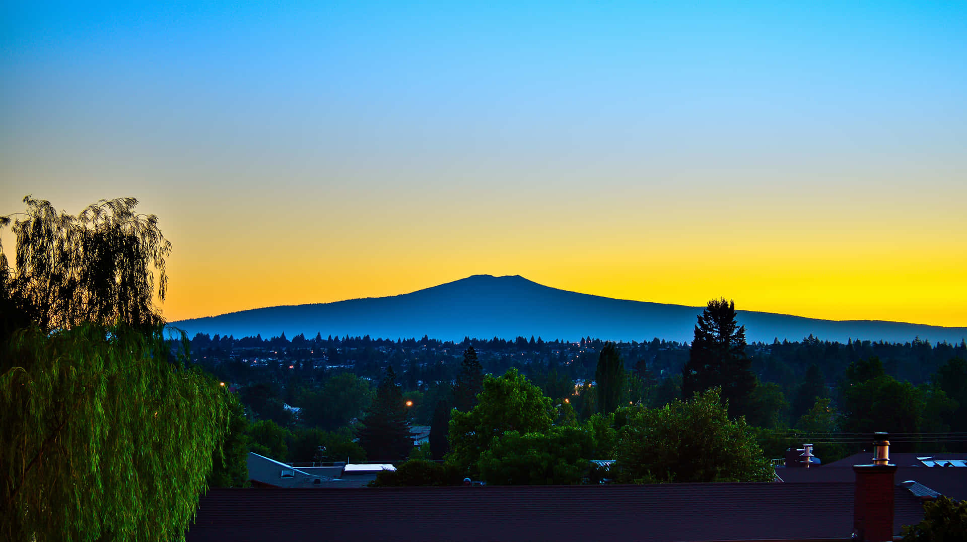 Horizon Silhouette with Vibrant Sky over Distant Peaks 壁紙 — 寧靜的日落將天空染成金黃色和藍色，遠處的山巒輪廓在郊區的屋頂和樹木上方升起。