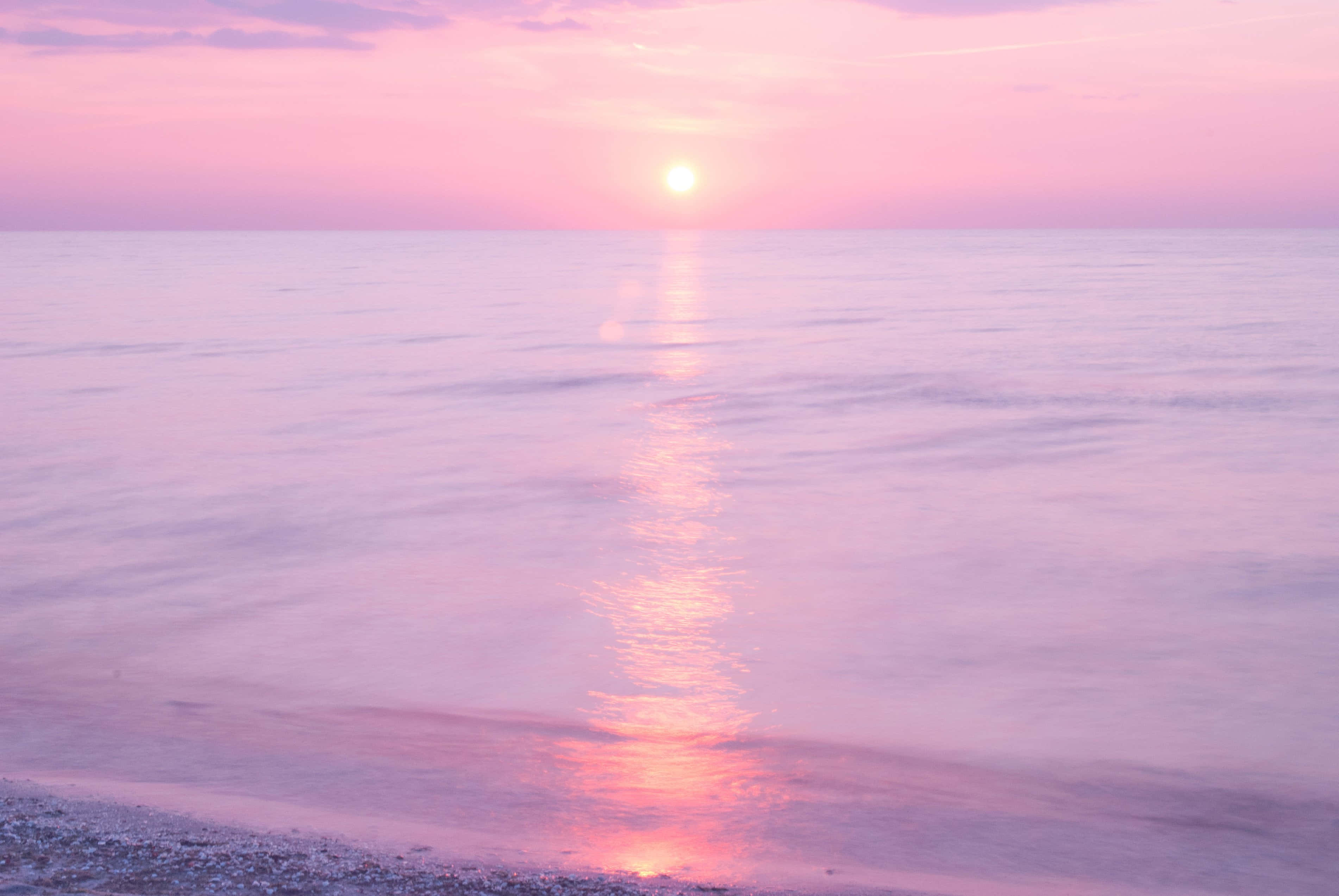 Pink Beach Sunset 桌布 壁紙 — 寧靜的日落映照在平靜的水面上，粉紅色和橙色的光芒在卵石灘附近的溫柔波浪上反射。