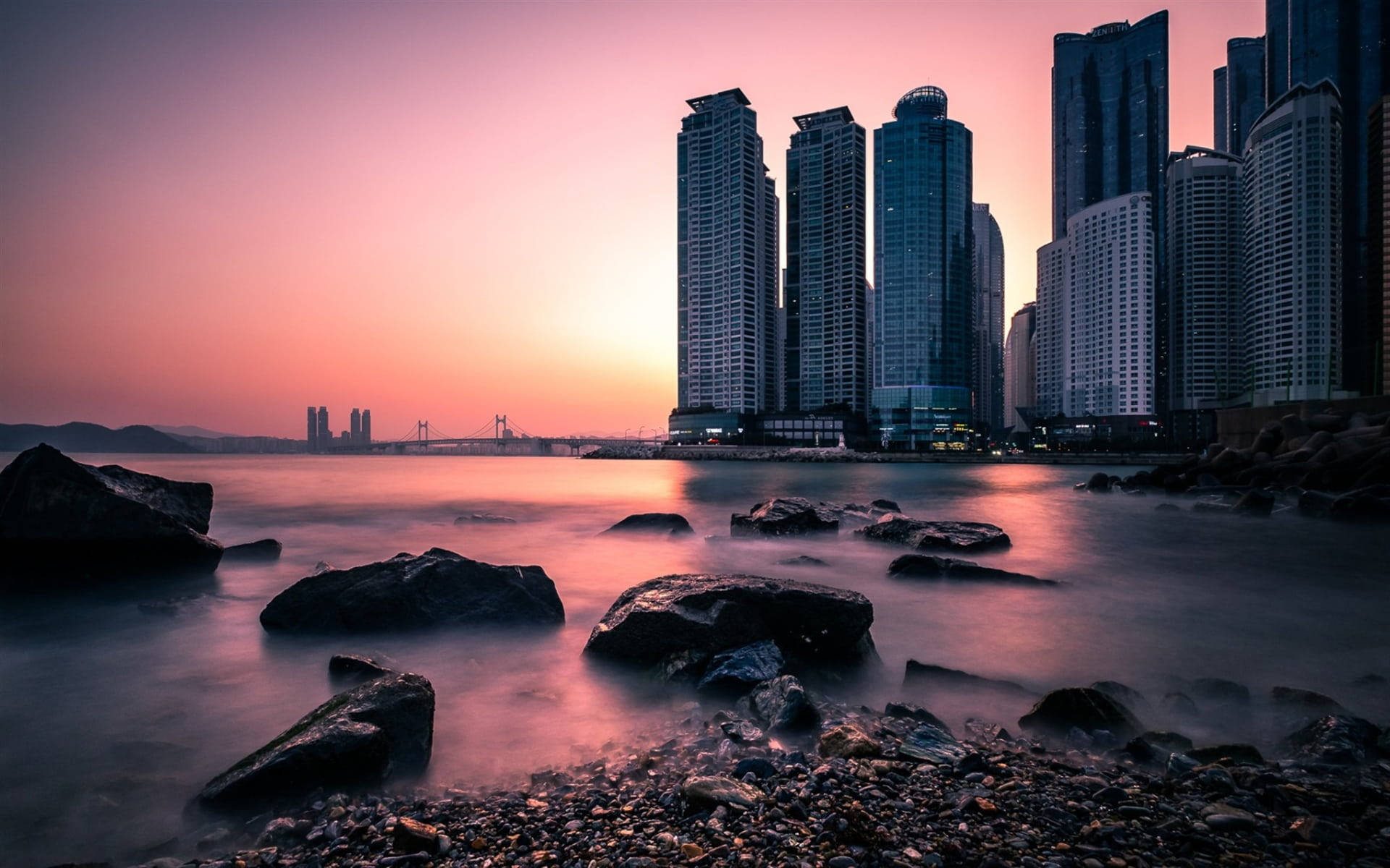 Busan 桌布 壁紙 — 寧靜的日落映照現代城市天際線，前景是平靜的水面和深色岩石。