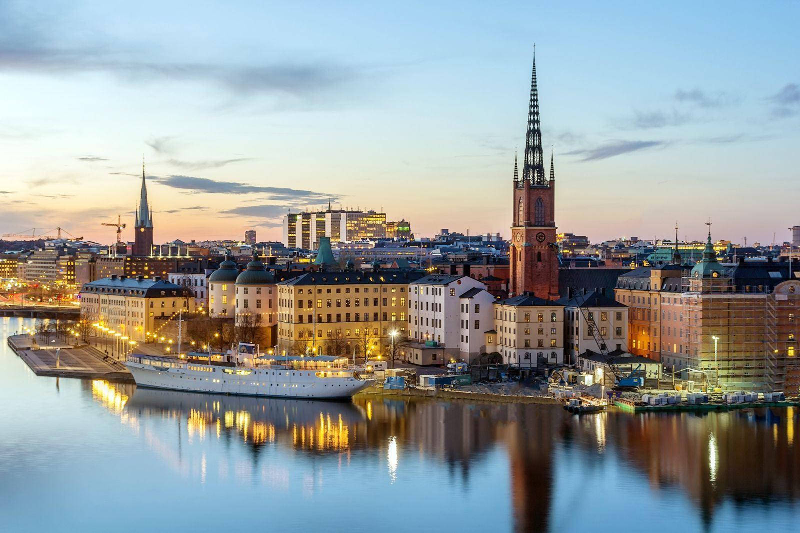Sweden 桌布 壁紙 — 寧靜的暮色城市景觀，歷史建築與白色船隻在平靜的水面上倒映，天空柔和湛藍。