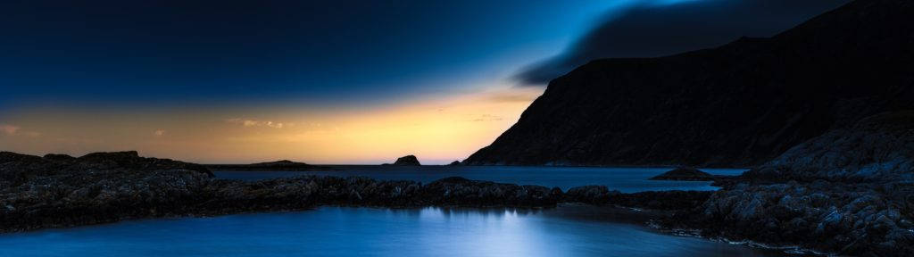 Cool 4K 桌布 壁紙 — 寧靜的暮色海景，平靜的藍色海水在黑暗的岩石海岸和剪影懸崖之間反射出金色的地平線。