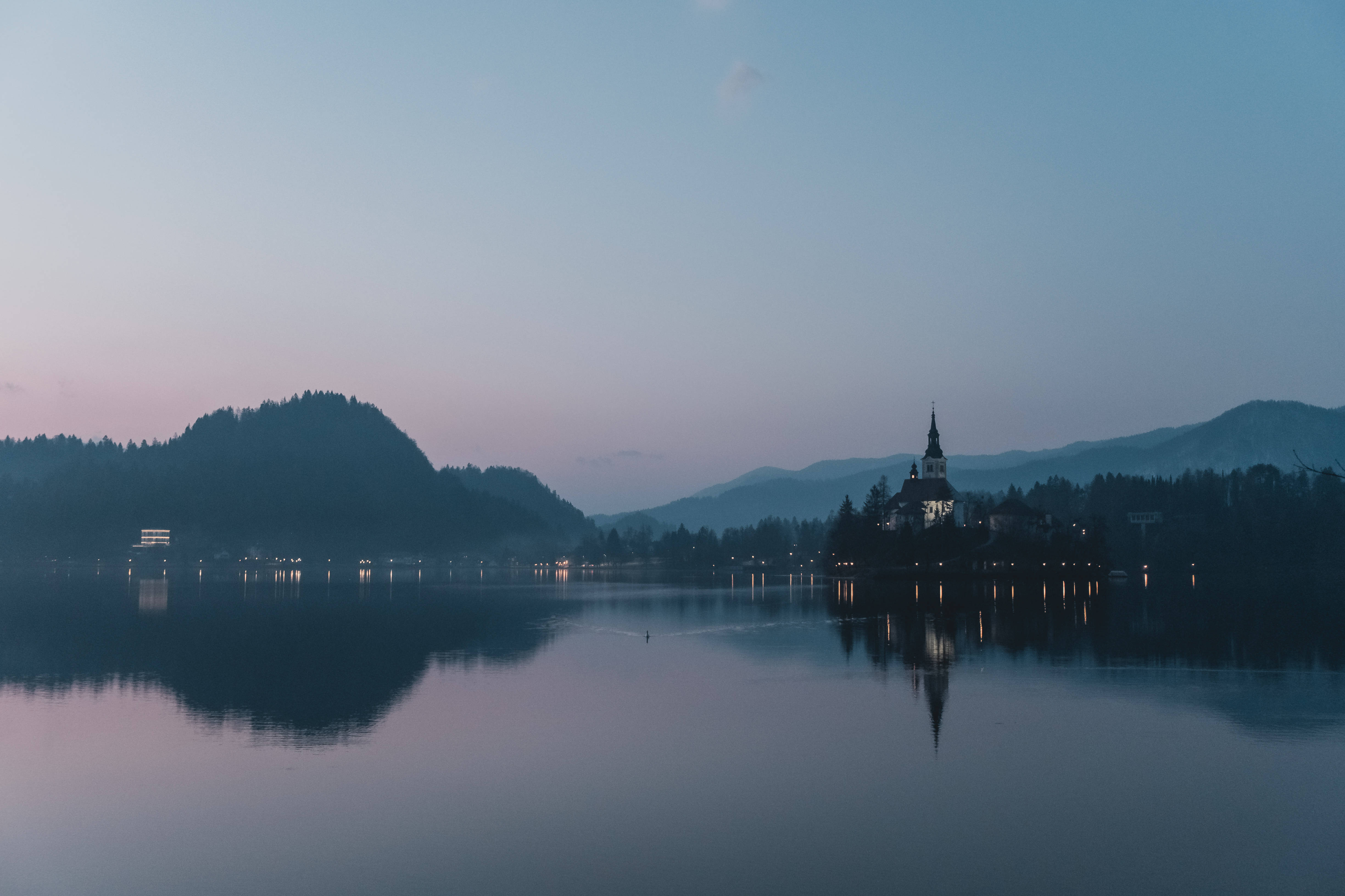 Slovenia 桌布 壁紙 — 寧靜的暮色湖泊在柔和的紫色和藍色天空下倒映著教堂和遠山。