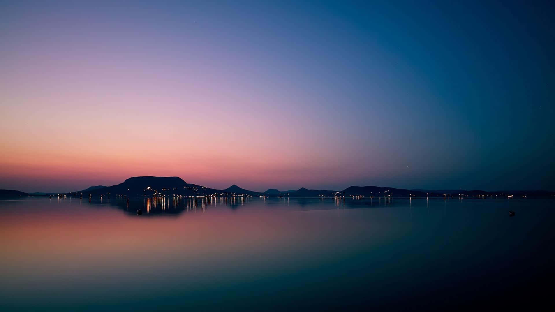 Apple Mac Desktop 桌布 壁紙 — 寧靜的暮色湖泊映照出紫色、粉紅色和深藍色的漸變天空，遠方有山丘和城市燈光。