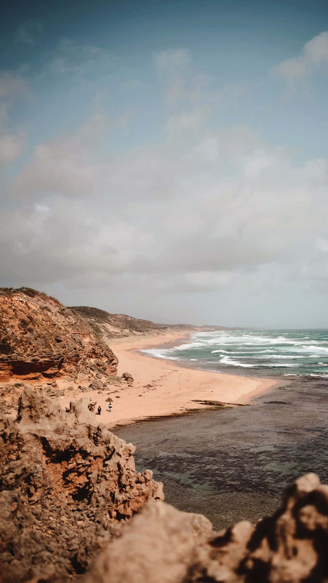 Beach Aesthetic Tumblr 桌布 壁紙 — 寧靜的海岸景色，崎嶇的懸崖俯瞰著沙灘，溫柔的海浪在多雲的天空下拍打著海岸。