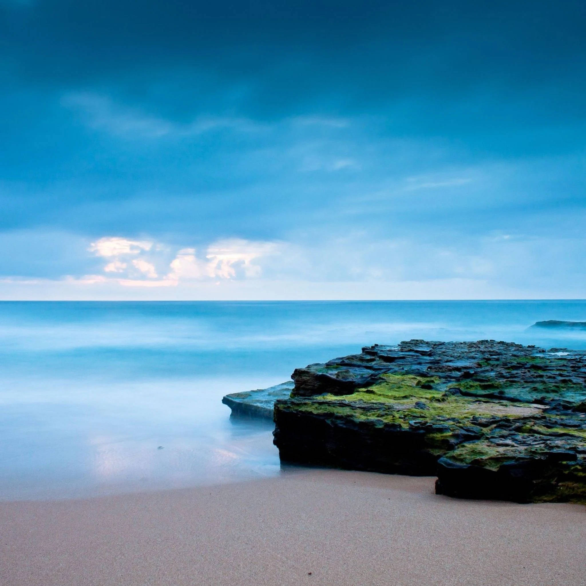 Beach Scene 桌布 壁紙 — 寧靜的海岸景色，平滑的碧綠海水流過長滿苔蘚的岩石，廣闊的藍天中飄著柔軟的白雲。