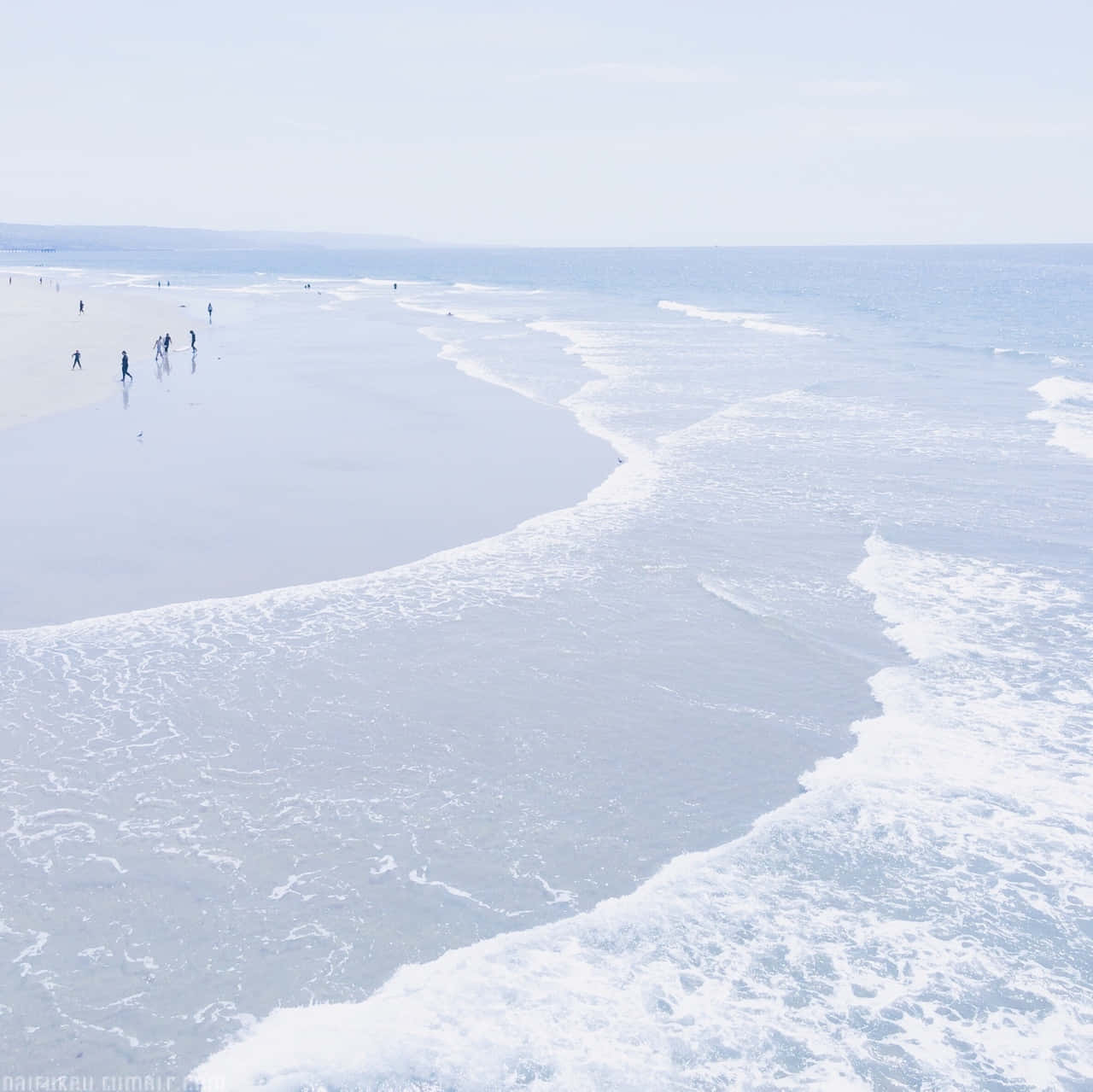 Ocean Beach 桌布 壁紙 — 寧靜的海岸景色，柔和的海浪拍打著寬闊的沙灘，淡藍色的天空下，遠處有人沿著海岸漫步。