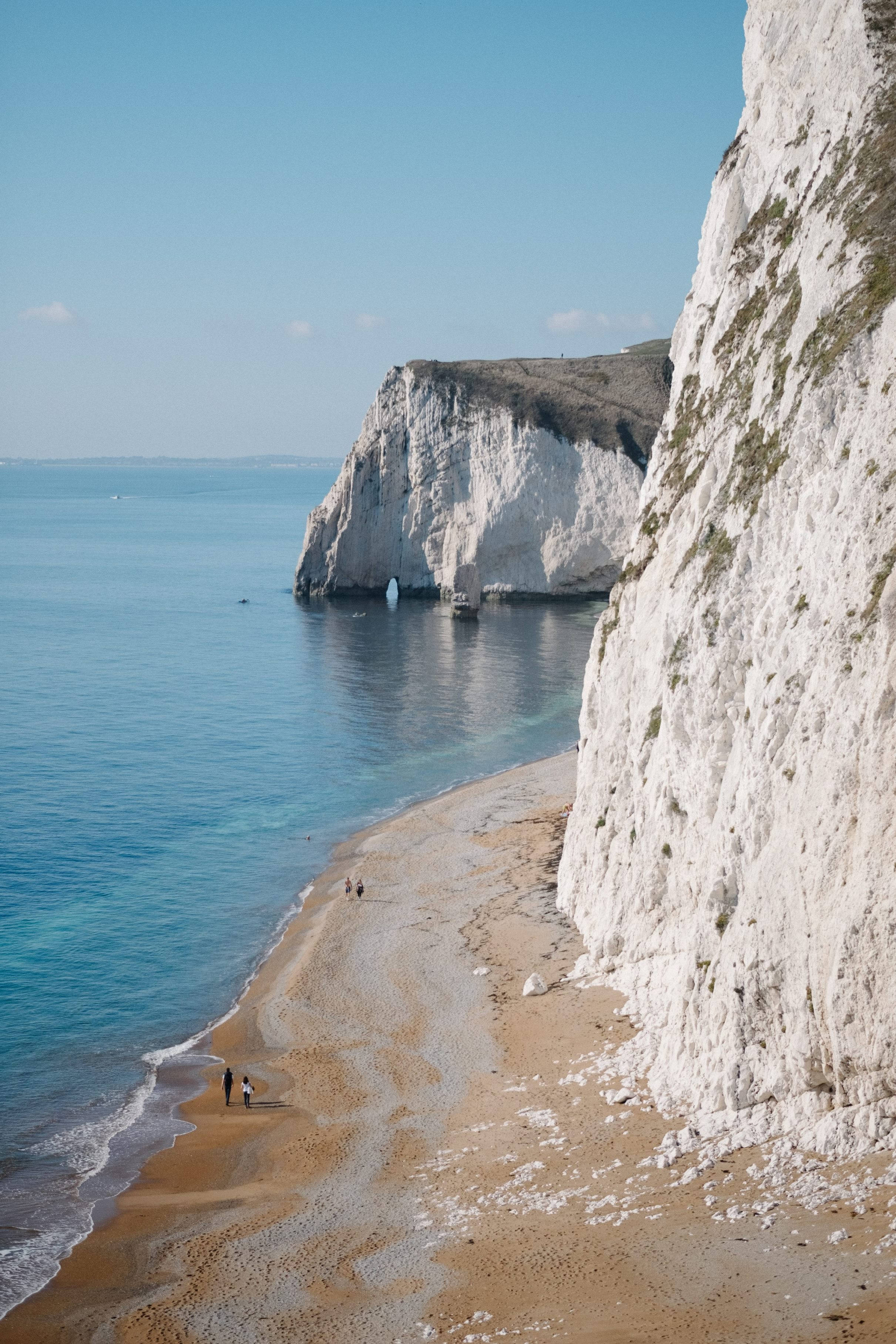 England 桌布 壁紙 — 寧靜的海岸景色，高聳的白色懸崖俯瞰著碧綠色的海洋和沙灘，沙灘上有細小的人影沿著海岸行走。