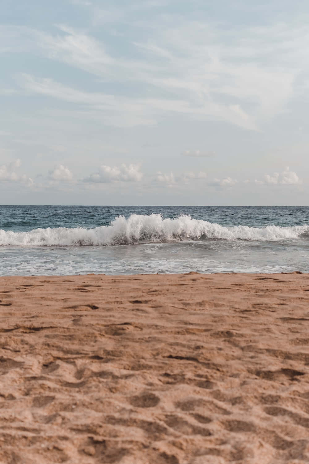 Minimalist Beach 桌布 壁紙 — 寧靜的海灘場景，前景有紋理沙灘，柔和的海浪在柔軟多雲的天空下拍打。
