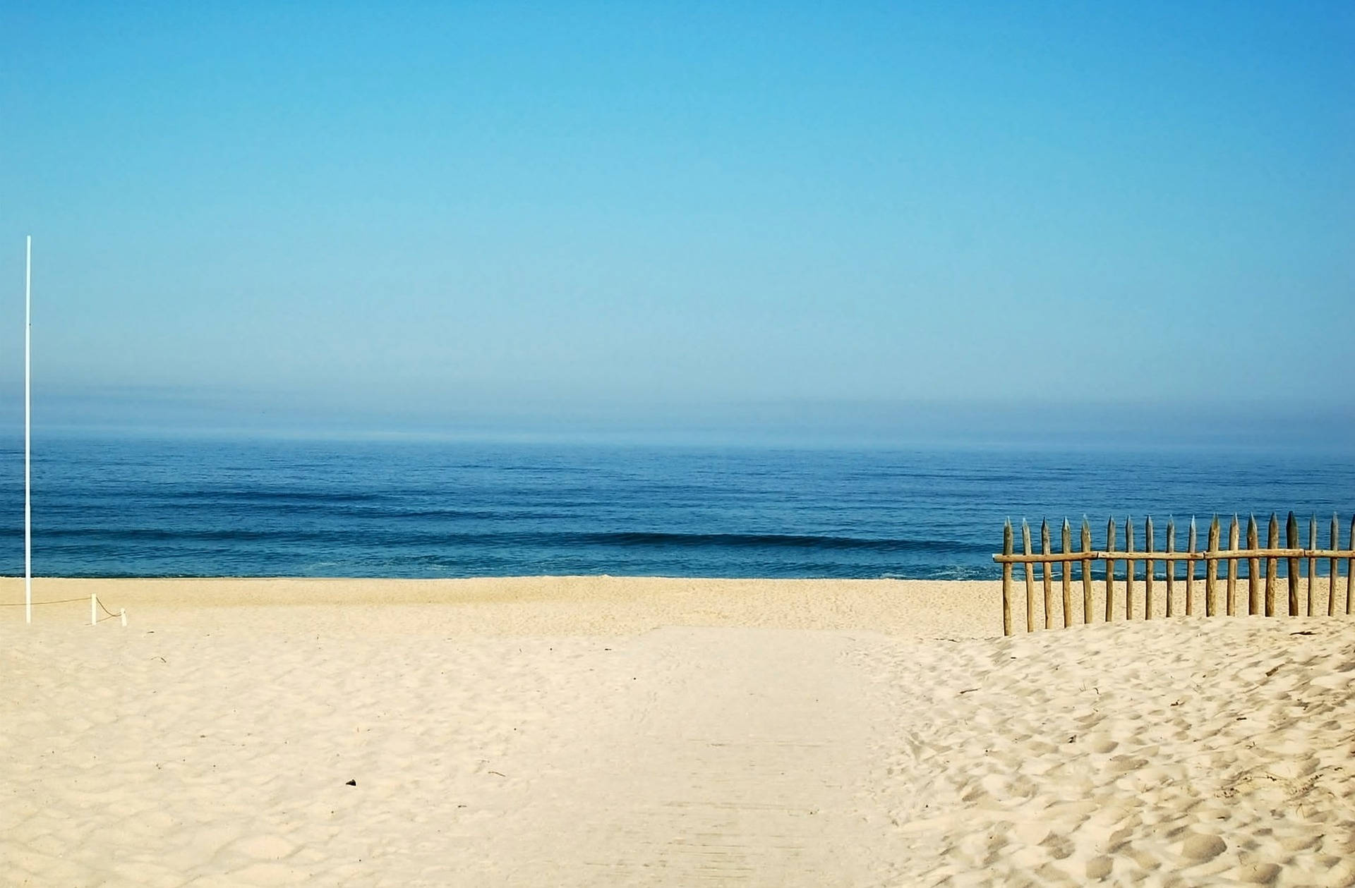 Beach Desktop 桌布 壁紙 — 寧靜的海灘場景，柔軟的白沙、平靜的藍色海洋，以及晴朗天空下的木柵欄。