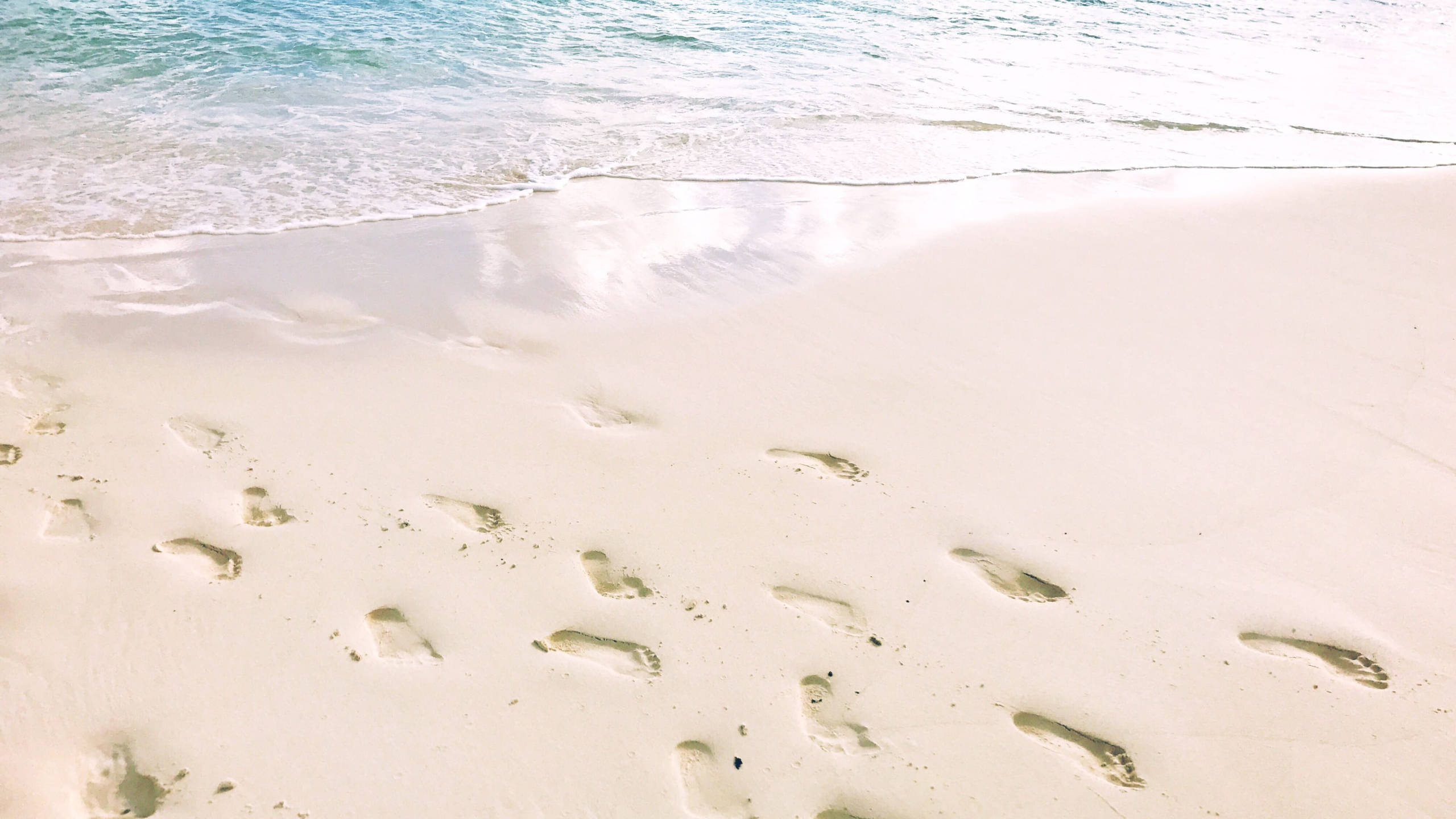 White Beach 桌布 壁紙 — 寧靜的海灘場景，溫柔的海浪拍打著純淨的白沙，沙灘上有一串腳印通往碧綠的海水。