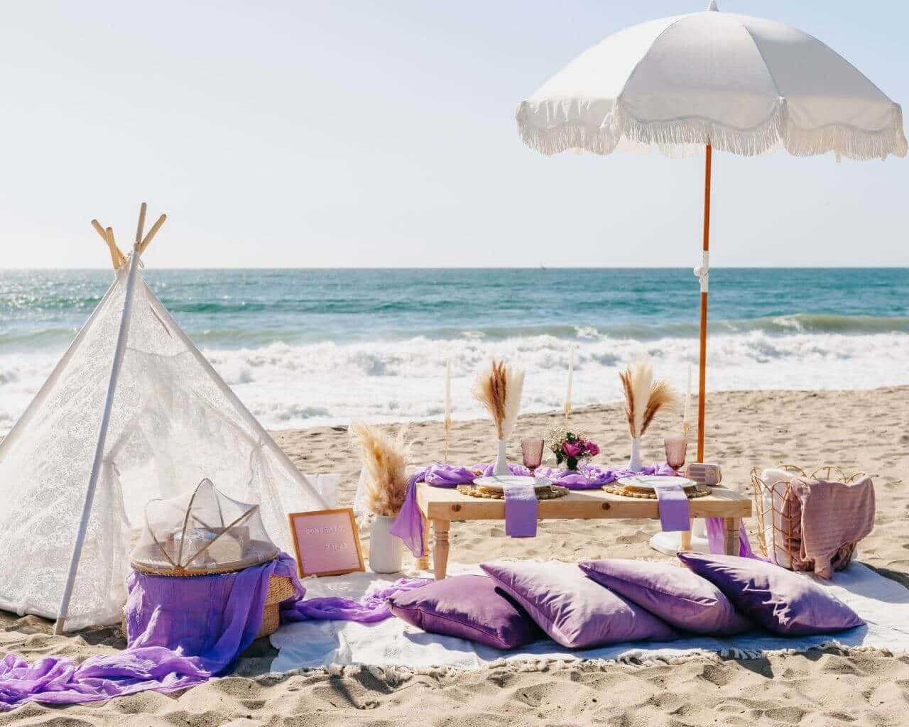 Beach Picnic 桌布 壁紙 — 寧靜的海灘野餐佈置，設有白色帳篷和紫色靠墊，位於海邊帶流蘇的遮陽傘下。