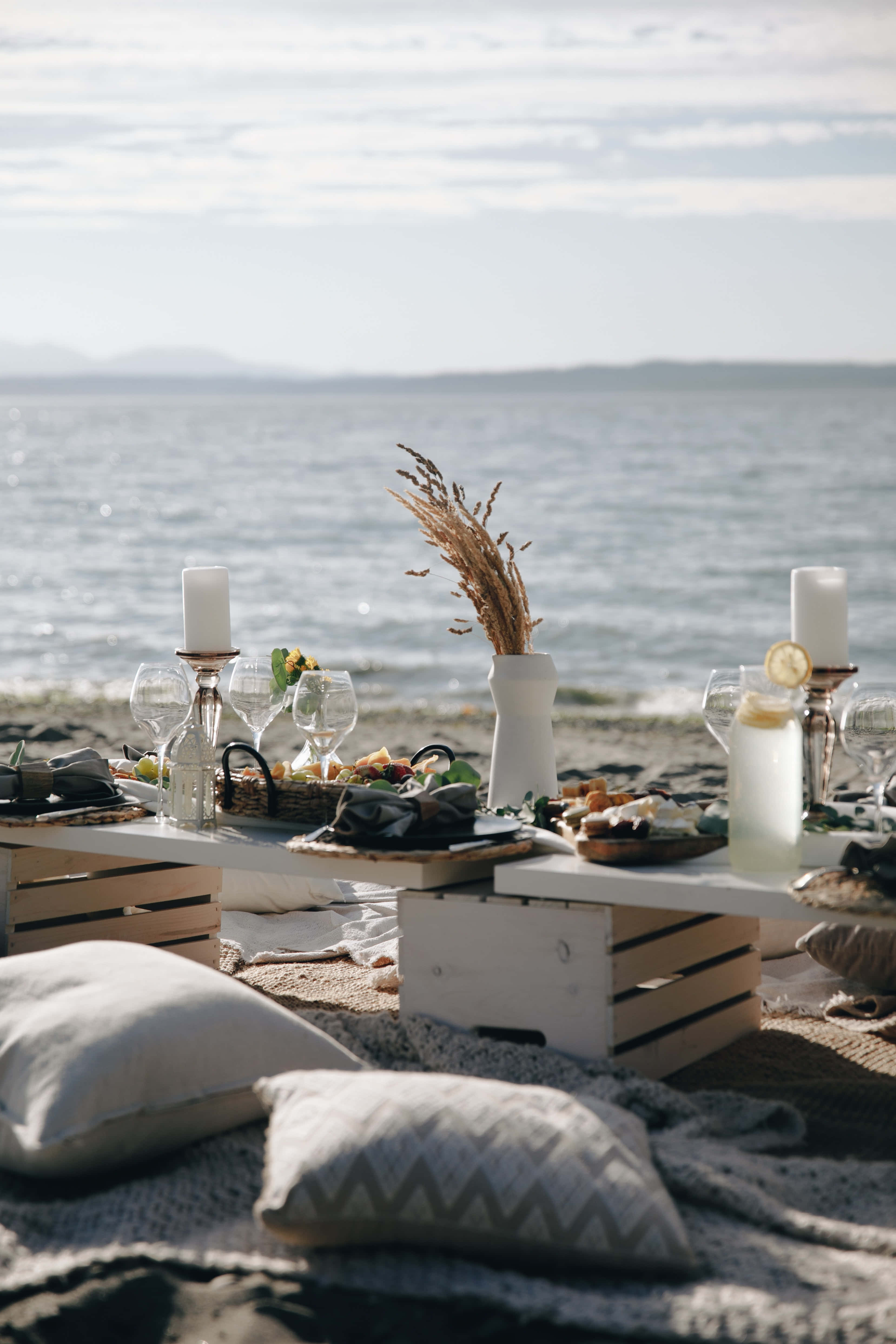 Beach Picnic 桌布 壁紙 — 寧靜的海灘野餐佈置，配有白色木箱、餐具和枕頭，在柔和日光下靠近平靜的海水。