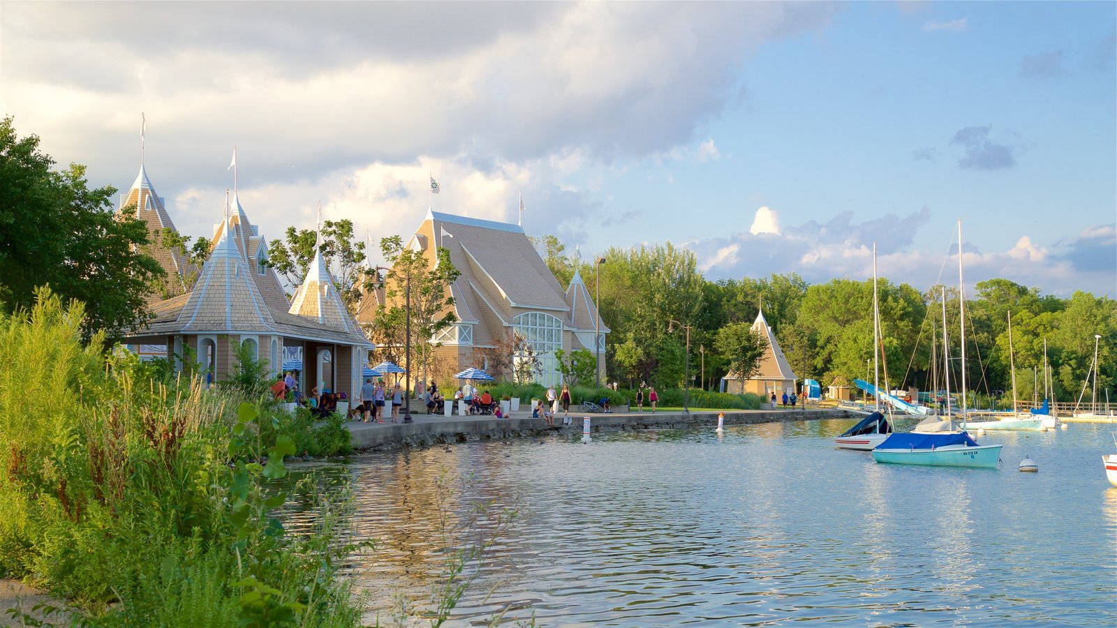 Minneapolis 桌布 壁紙 — 寧靜的湖邊景色，擁有獨特的尖頂建築、停泊的帆船，以及在部分多雲的天空下茂密的綠植。