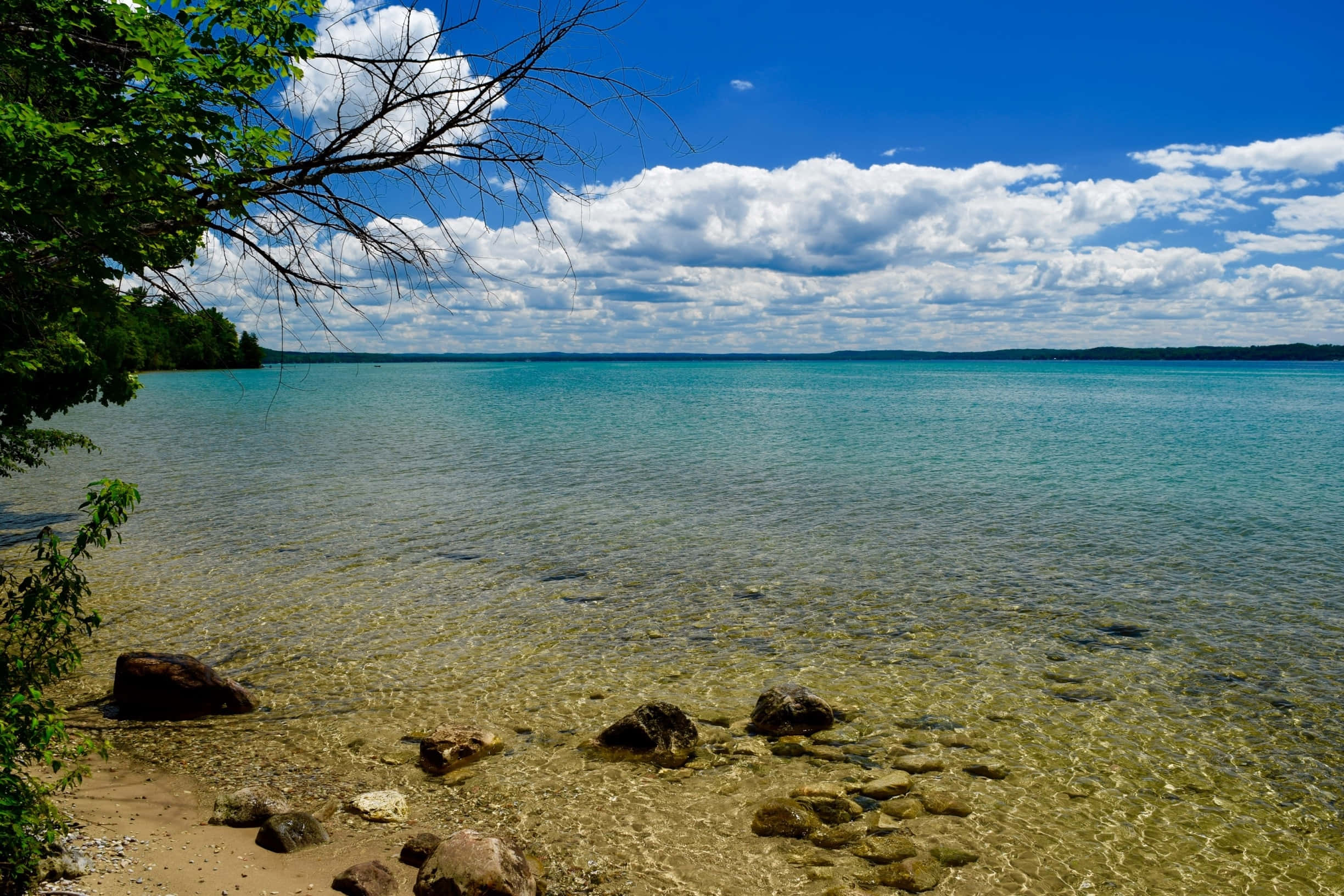 Torch Lake 桌布 壁紙 — 寧靜的湖邊景色，清澈的藍綠色淺水靠近岩石海岸，在明亮的藍天下有蓬鬆的白雲。