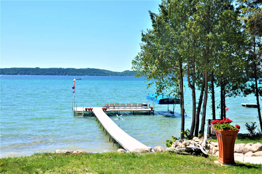 Torch Lake 桌布 壁紙 — 寧靜的湖邊碼頭延伸至碧綠色的水中，天空清澈湛藍，附近有茂密的綠樹和一盆紅花。