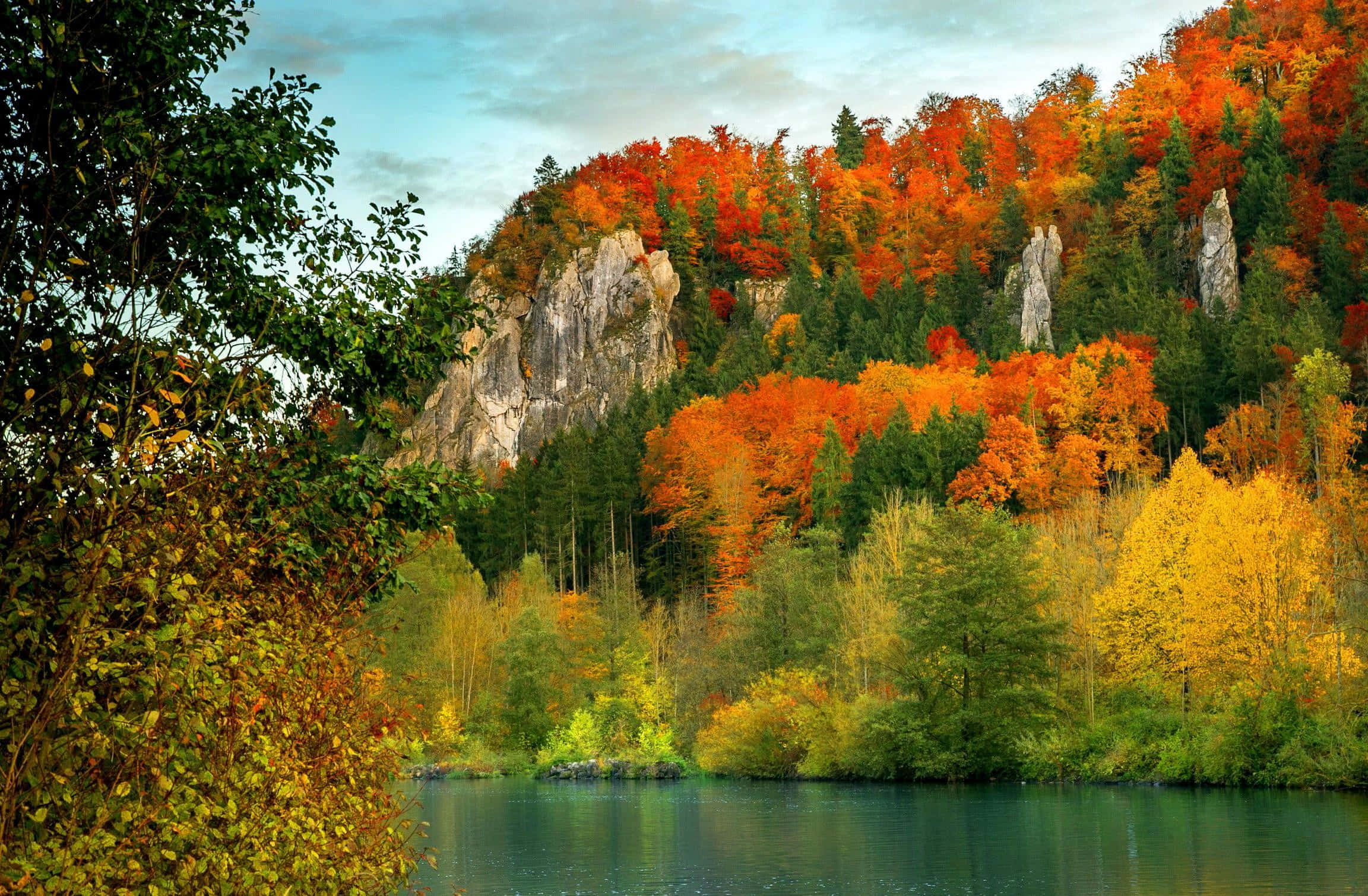 Fall Mountains 桌布 壁紙 — 寧靜的湖面倒映著充滿活力的秋日森林，火紅、橙黃和金色的樹木依附在岩石峭壁上，襯著柔和的藍天。